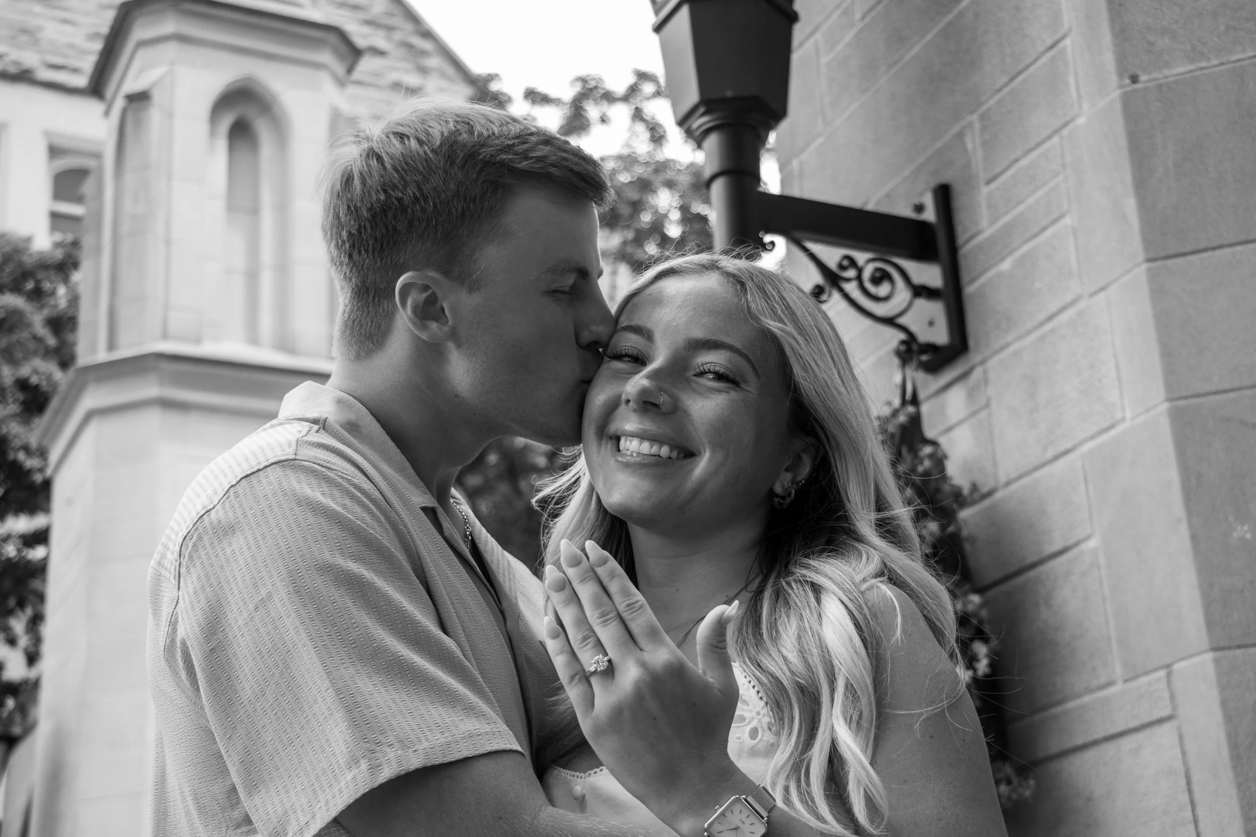 A black and white photo of a young man kissing a young woman on the forehead outside a building. The woman is smiling with her eyes closed, holding her hands together near her chest. The man is wearing a light-colored shirt, and the woman has light-c