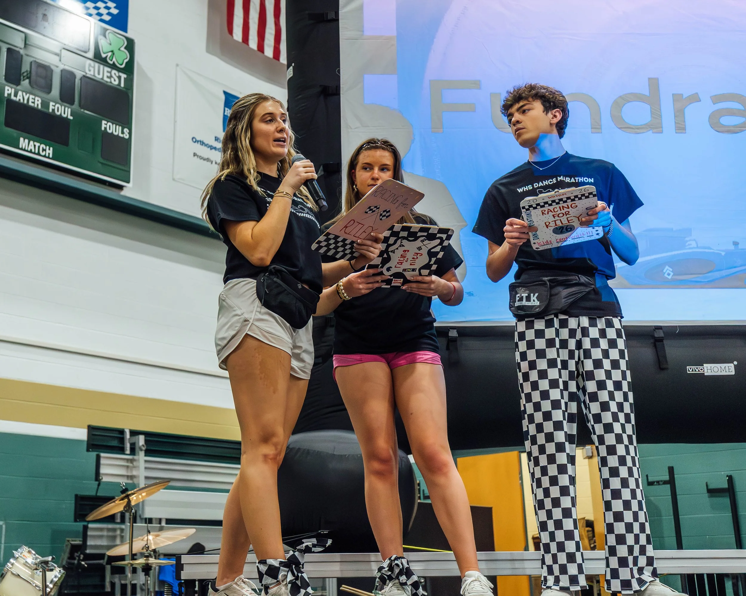 Three teenagers standing together in a gymnasium or sports facility, with the girl on the left holding a microphone and reading from a paper, the girl in the middle looking at her, and the boy on the right appearing to listen. They are dressed casual