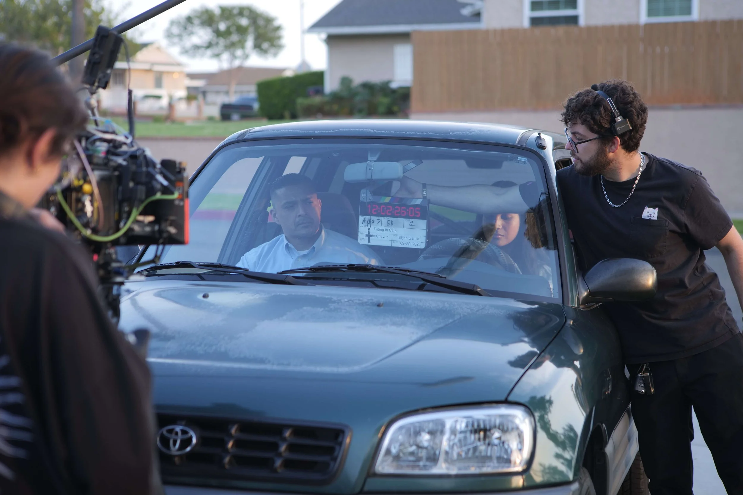 On a film set, a camera operator films a scene with two actors inside a green car. A man with curly hair and glasses leans over the car window speaking to the actors, while another crew member with headphones observes from the side.