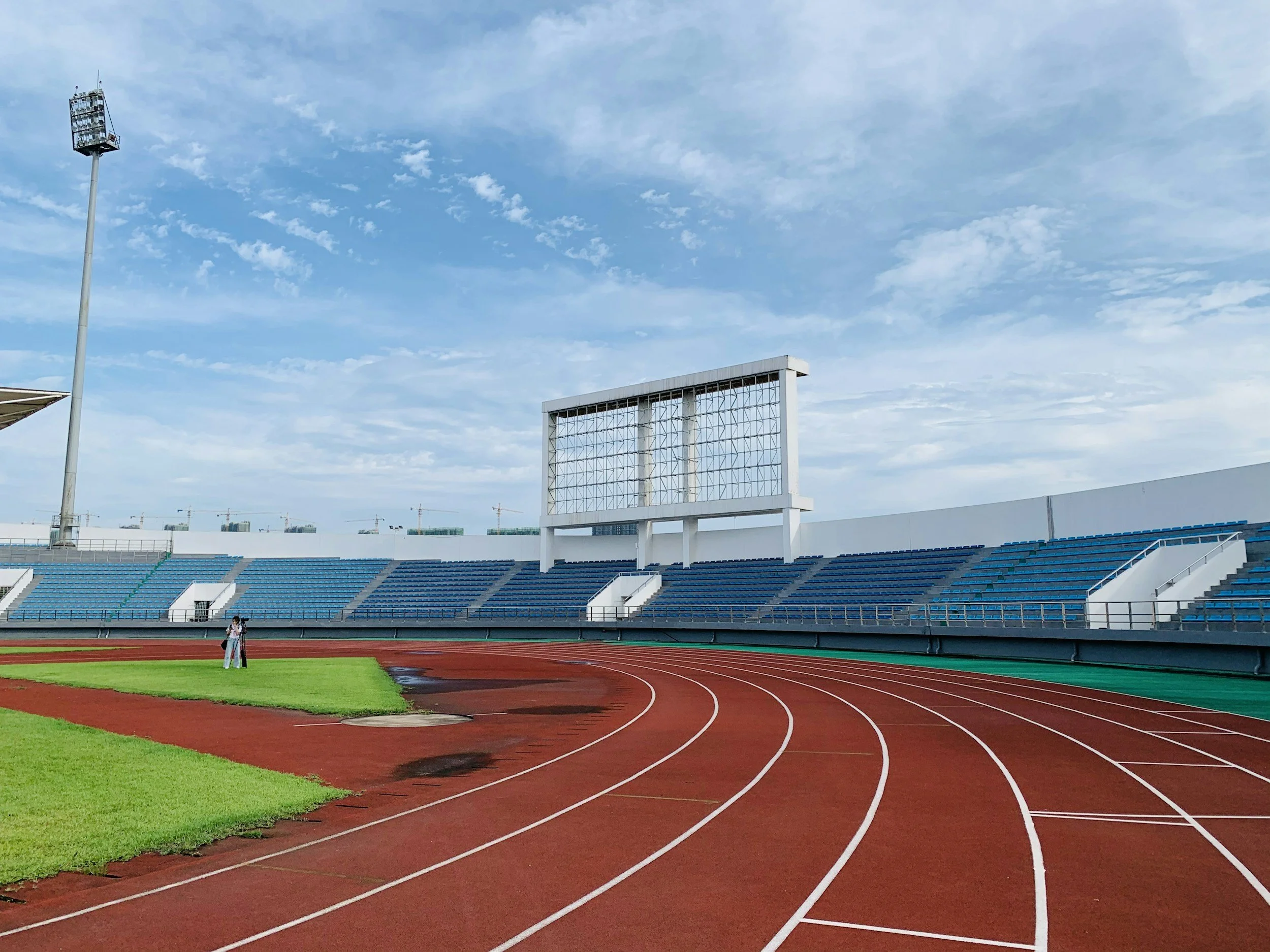 Empty outdoor running track at a sports stadium under partly cloudy sky with two people in the distance near the grass field.