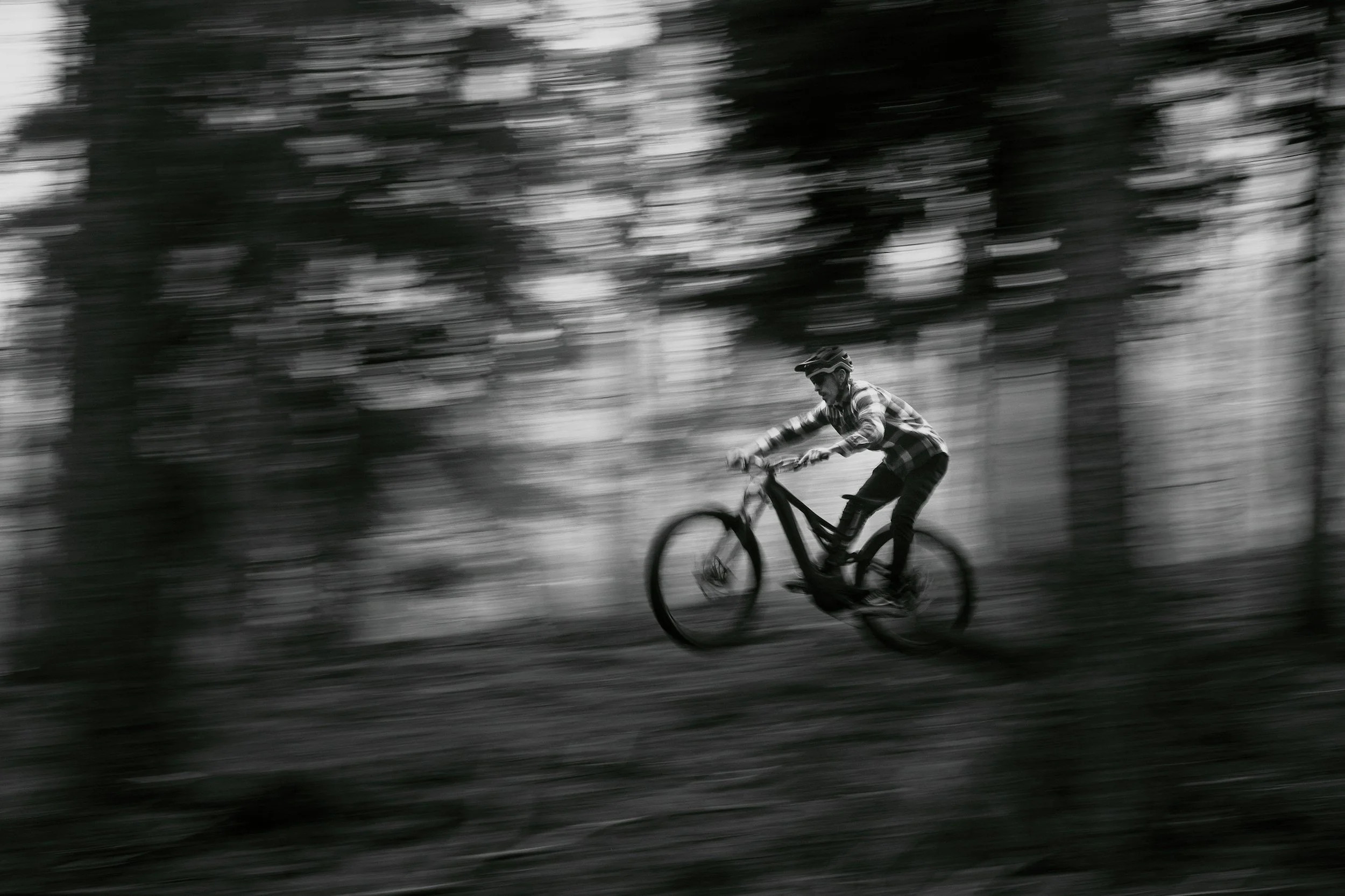 A person riding a mountain bike through a wooded area during daytime, captured with motion blur in black and white.