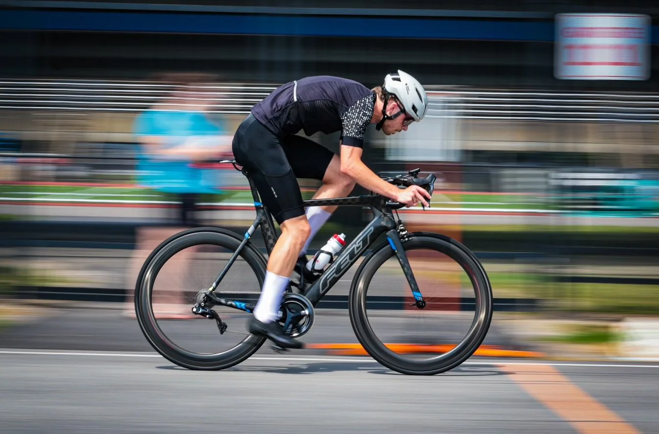 A cyclist wearing a helmet and sportswear riding a black and blue road bike at high speed on a city street, with motion blur in the background.