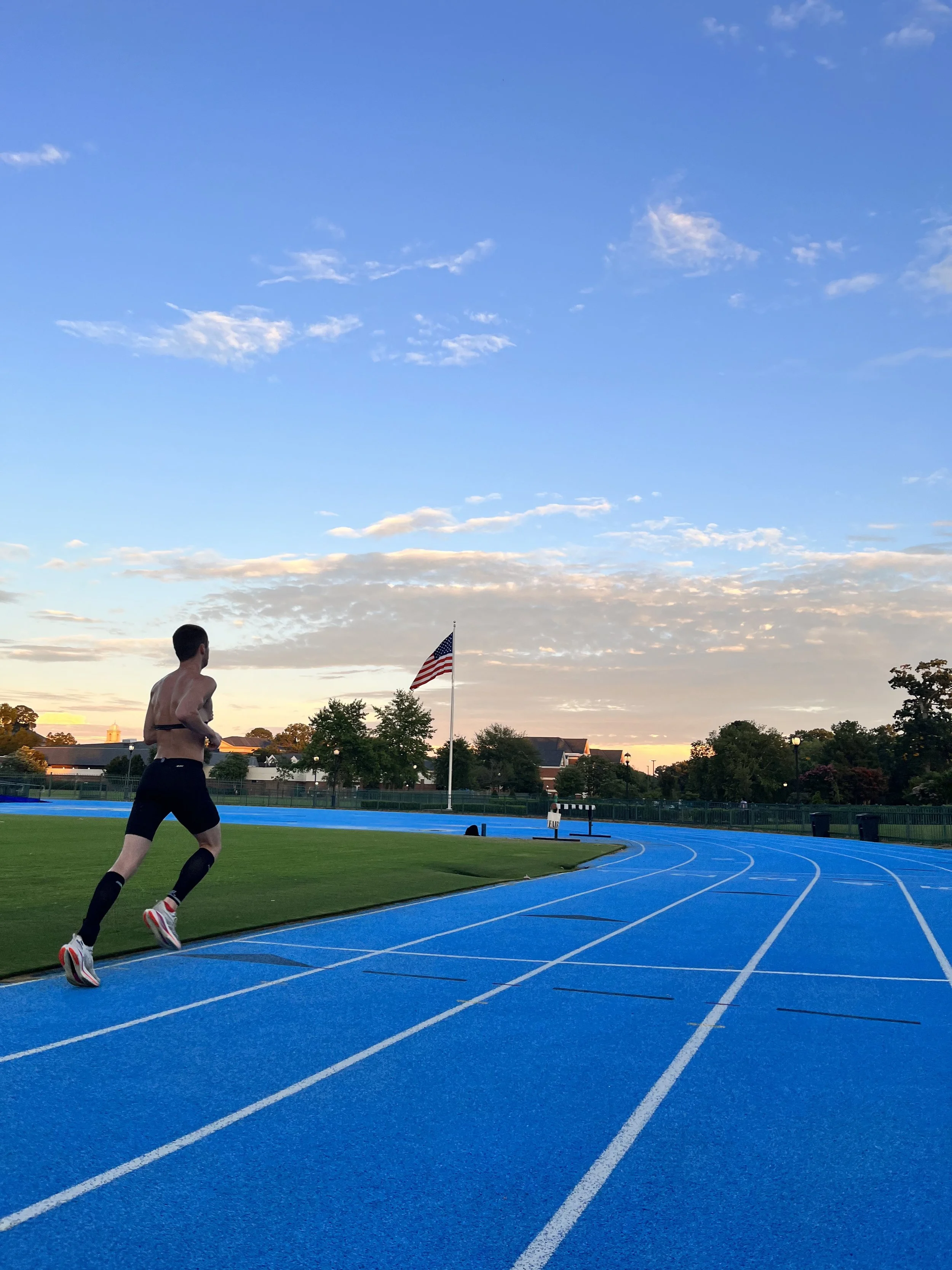 A male runner on a blue outdoor track during sunset, with an American flag and trees in the background.