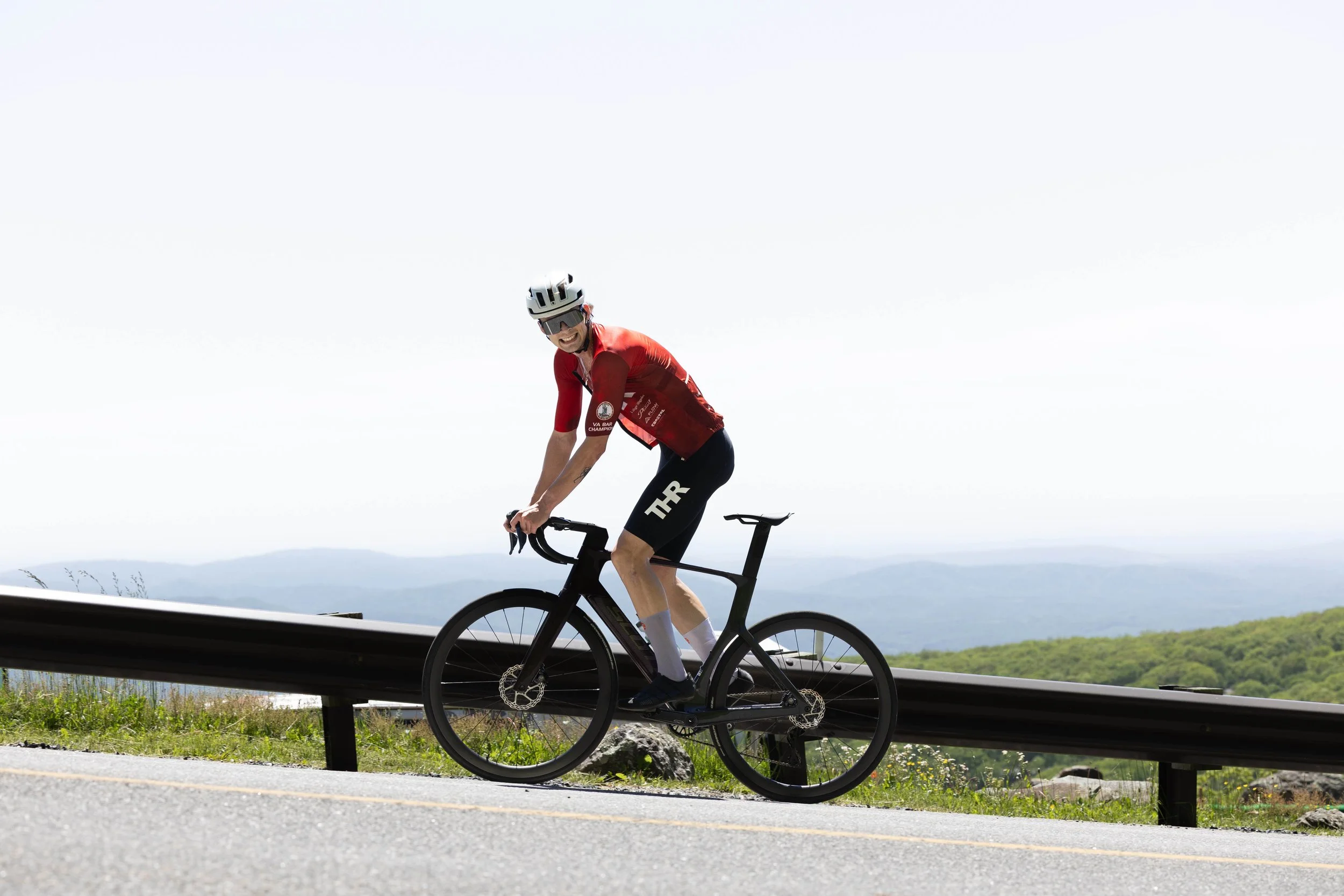 Cyclist wearing a red jersey and black shorts riding a black road bike on a mountain road with a guardrail, against a backdrop of rolling hills and a cloudy sky.