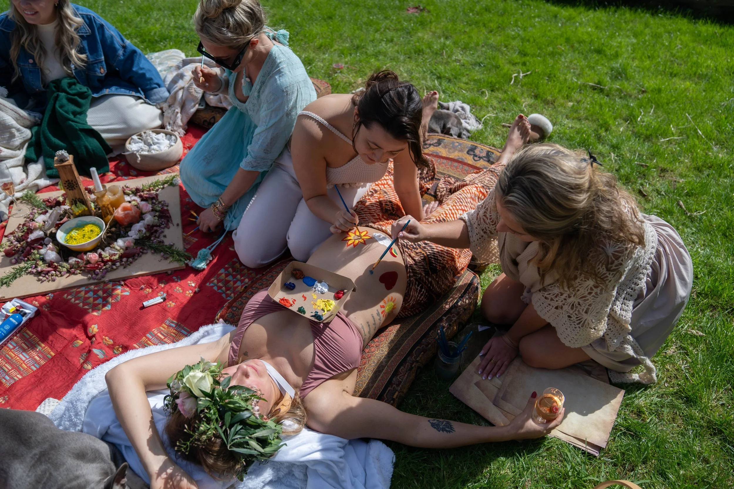 A group of women having a bohemian-style outdoor gathering on a grassy field, with one woman lying down with a flower crown, while others paint body art on her, surrounded by colorful decorations, paints, and objects.