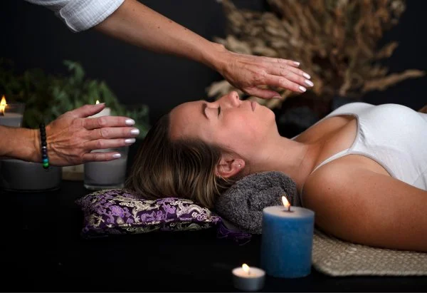A dimly lit room decorated with Halloween decorations including hanging paper skulls and banners, with yellow string lights creating streaks of light. Two women are on a bed, one lying down and the other sitting beside her, in a cozy, decorated space.