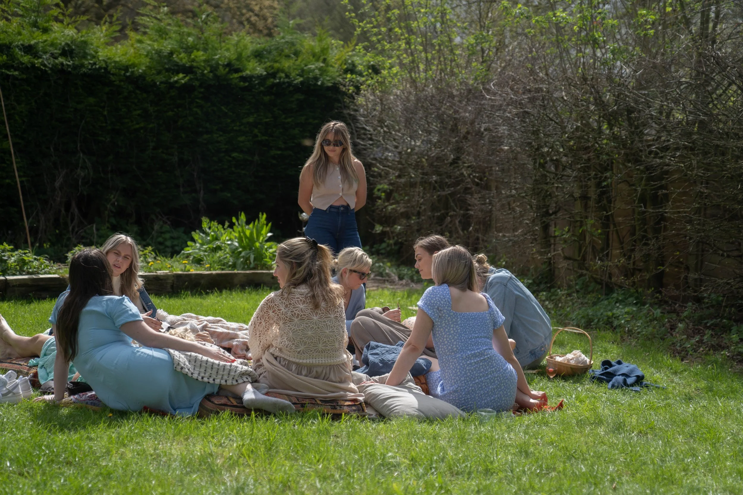Group of women sitting on a blanket in a lush green garden, some engaging in conversation and one standing nearby.