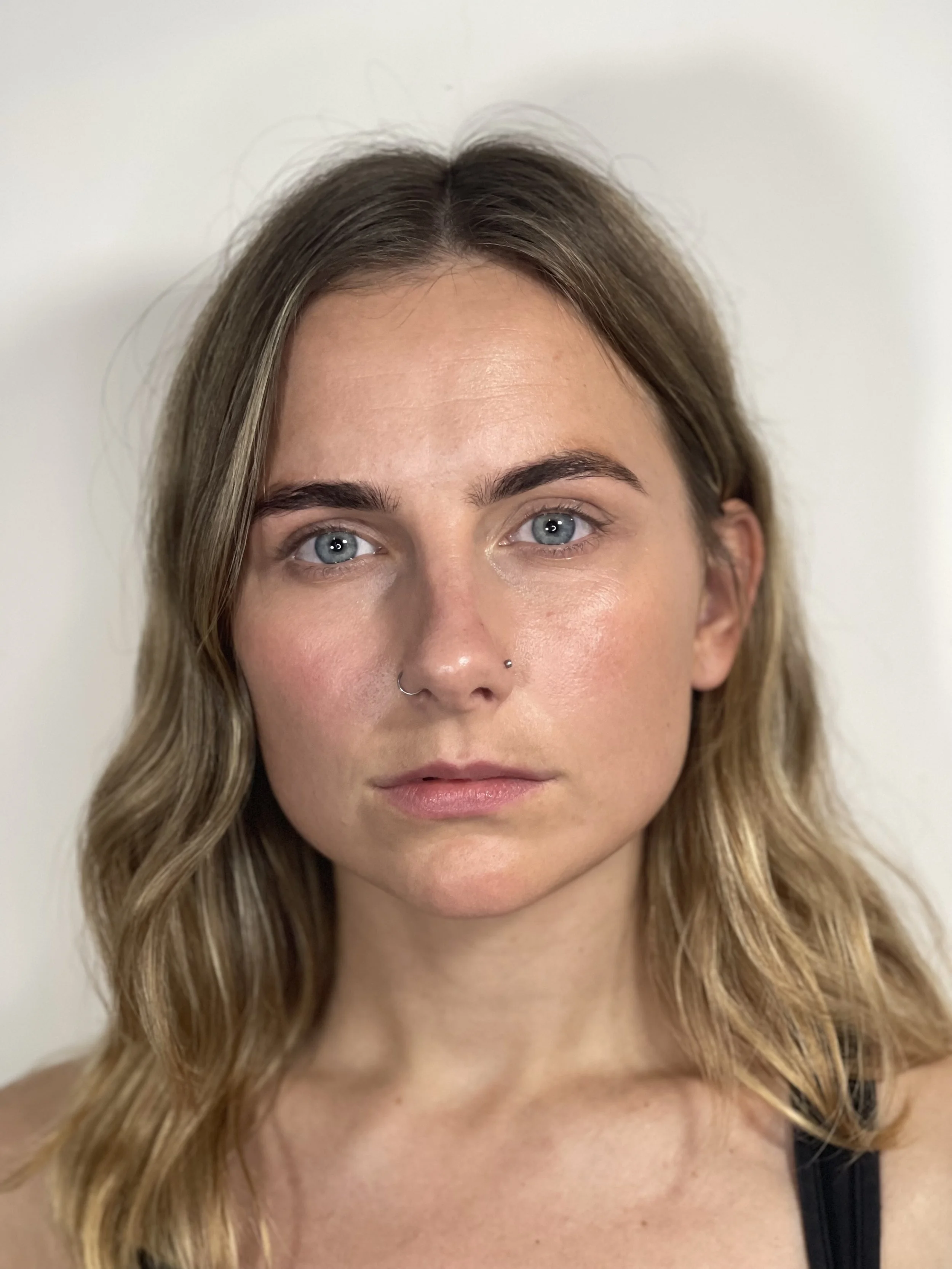 A close-up portrait of a woman with shoulder-length wavy hair, blue eyes, and a nose piercing, looking directly at the camera with a neutral expression.