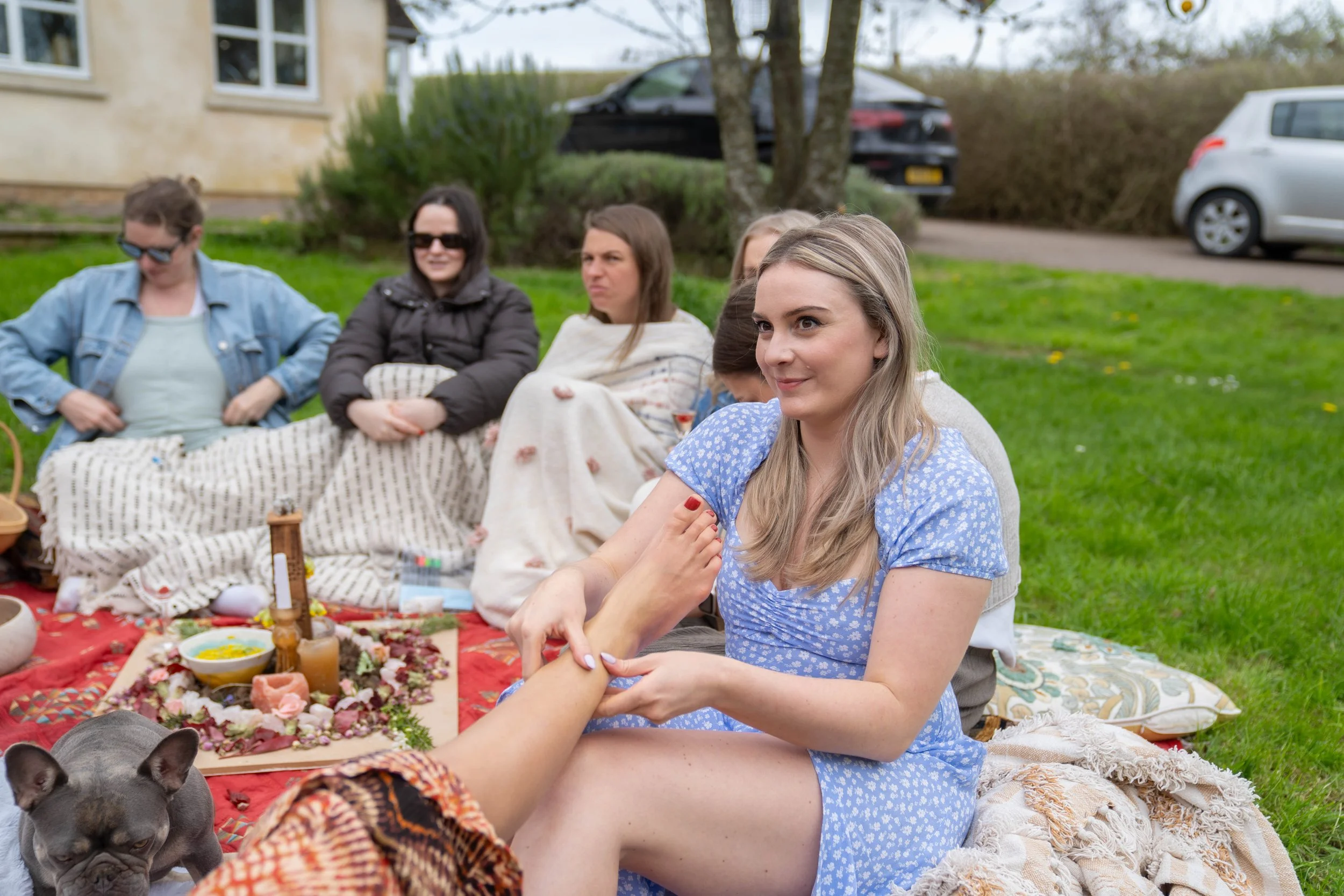 A group of women having a picnic outdoors, with one woman smiling and holding hands with someone off-camera. They are sitting on blankets with food and drinks, with cars and trees in the background.