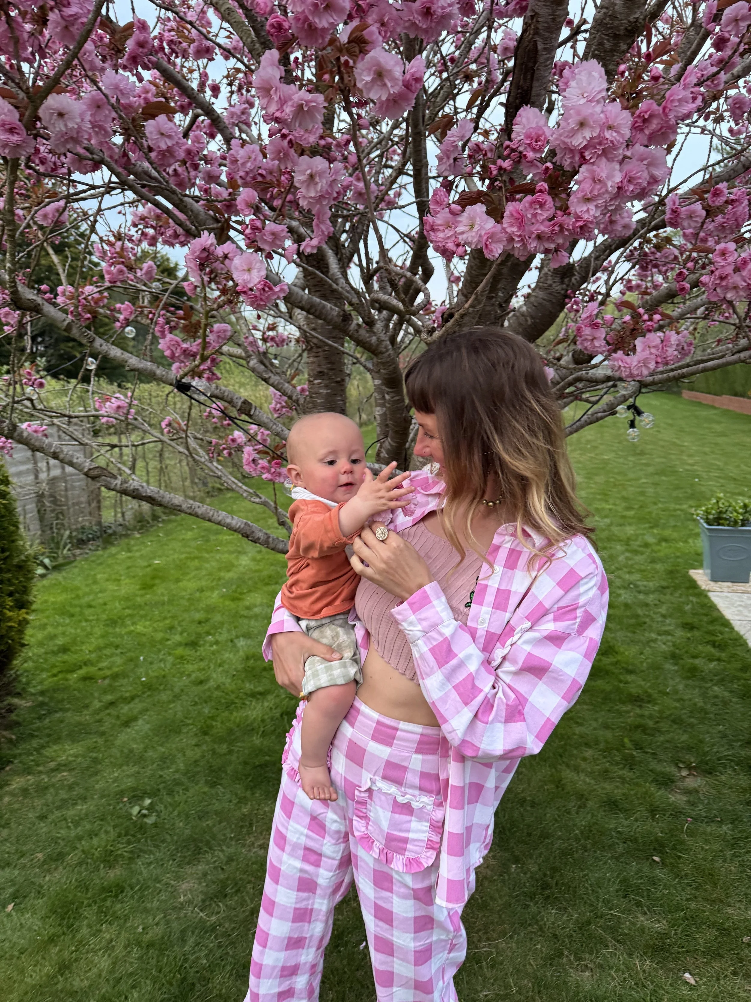 A woman wearing pink and white checkered pajamas holding a baby in front of a cherry blossom tree in a garden.