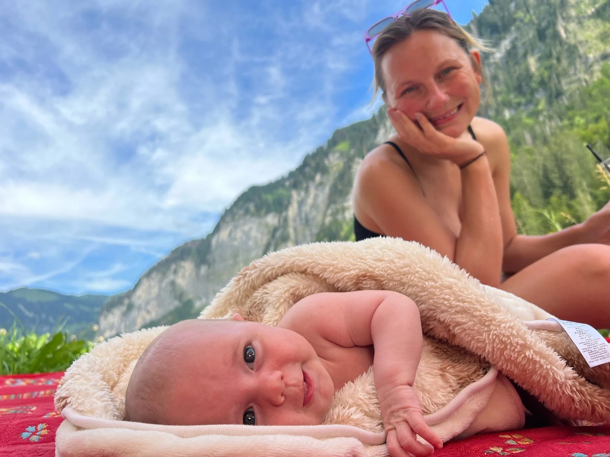 A woman and a baby lying outdoors on a blanket, with mountains and a blue sky in the background. The woman is smiling and resting her face on her hand, while the baby looks happy and relaxed under a soft blanket.