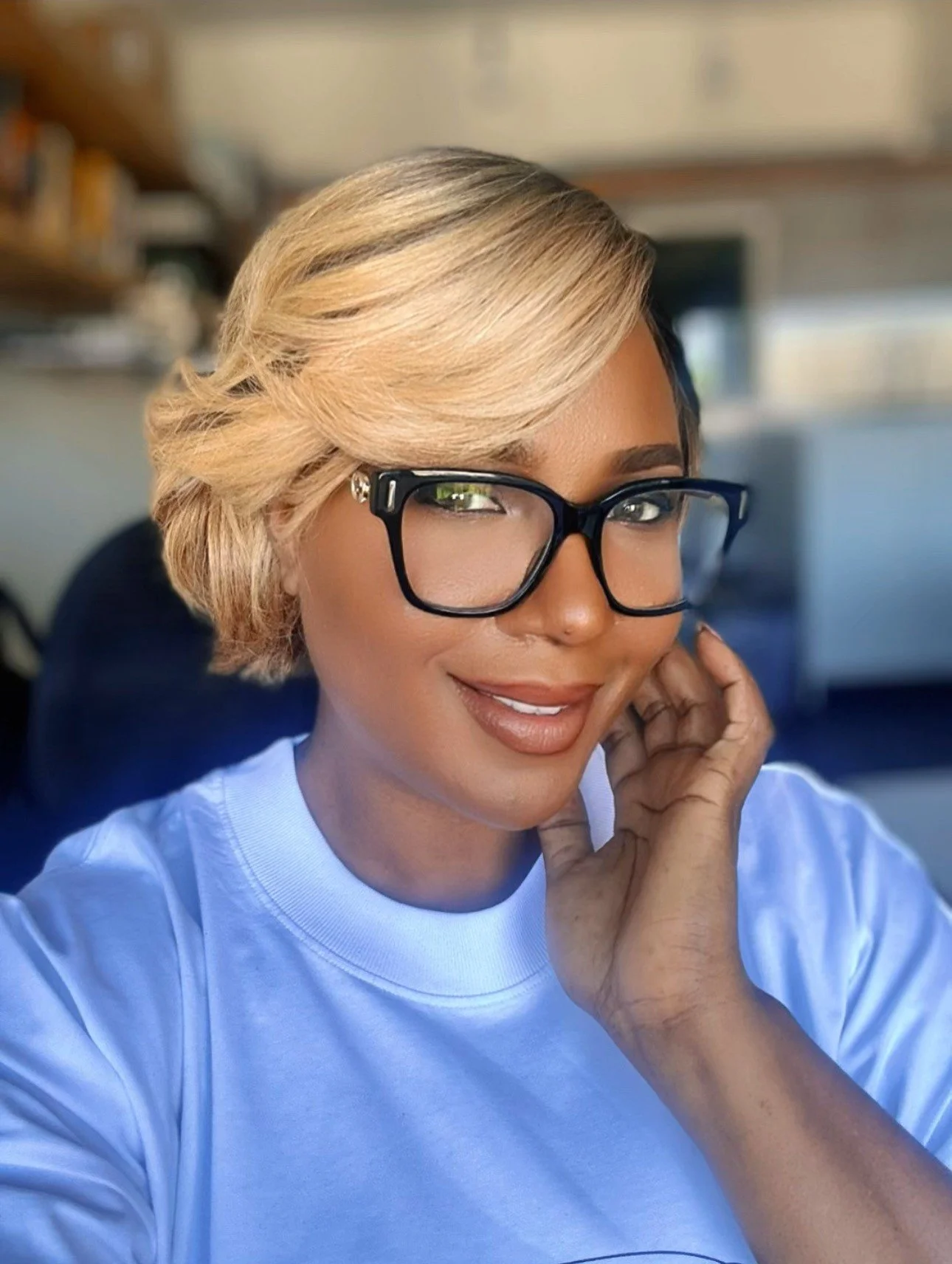 A woman with blonde hair, wearing glasses, smiling, touching her face with her right hand, indoors with a blurred background.