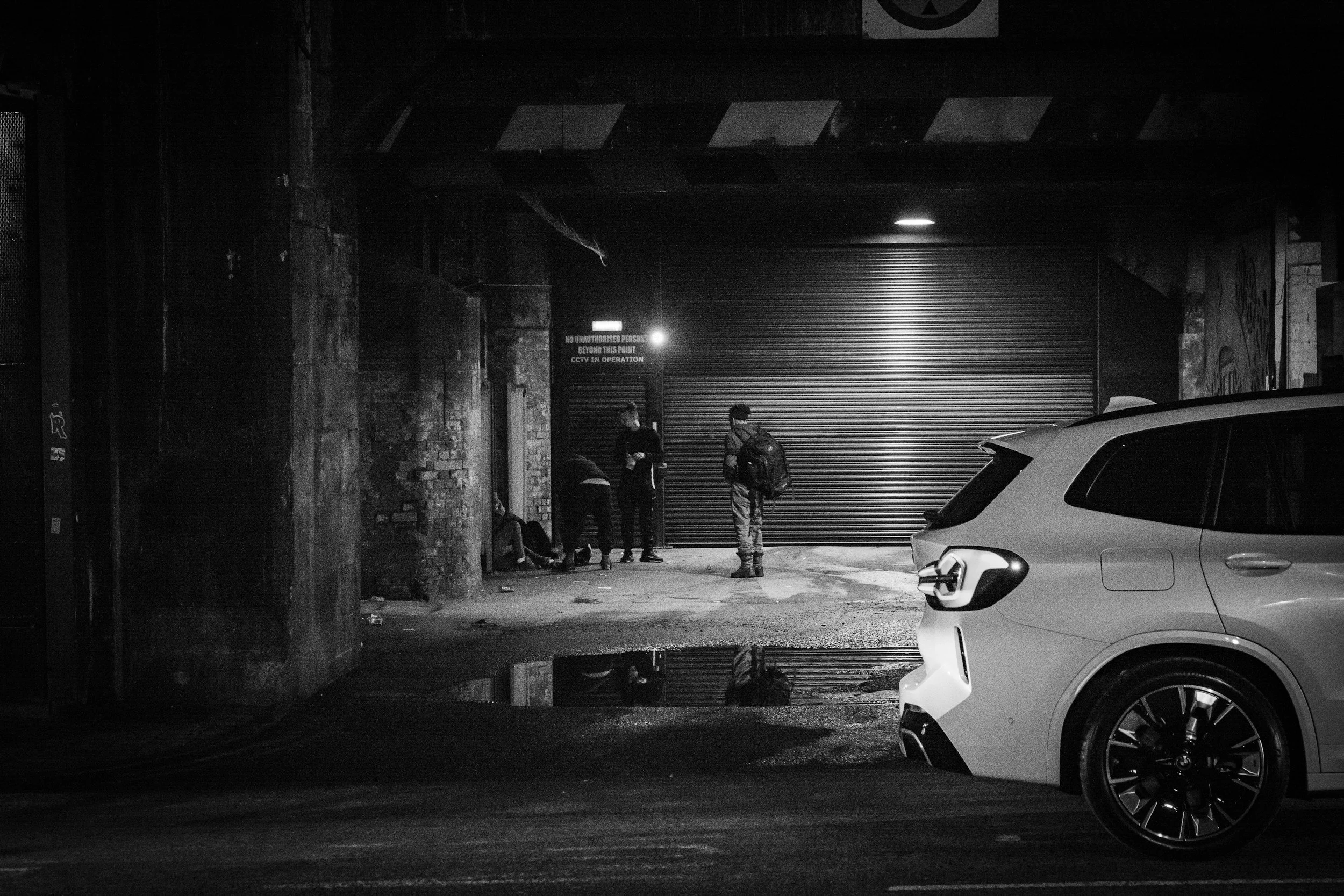 A black and white photo of a street scene at night showing a white SUV parked in the foreground, a group of four people gathered near a closed rolling metal door, with graffiti on the brick walls behind them. Street lighting and a sign are visible in the background.
