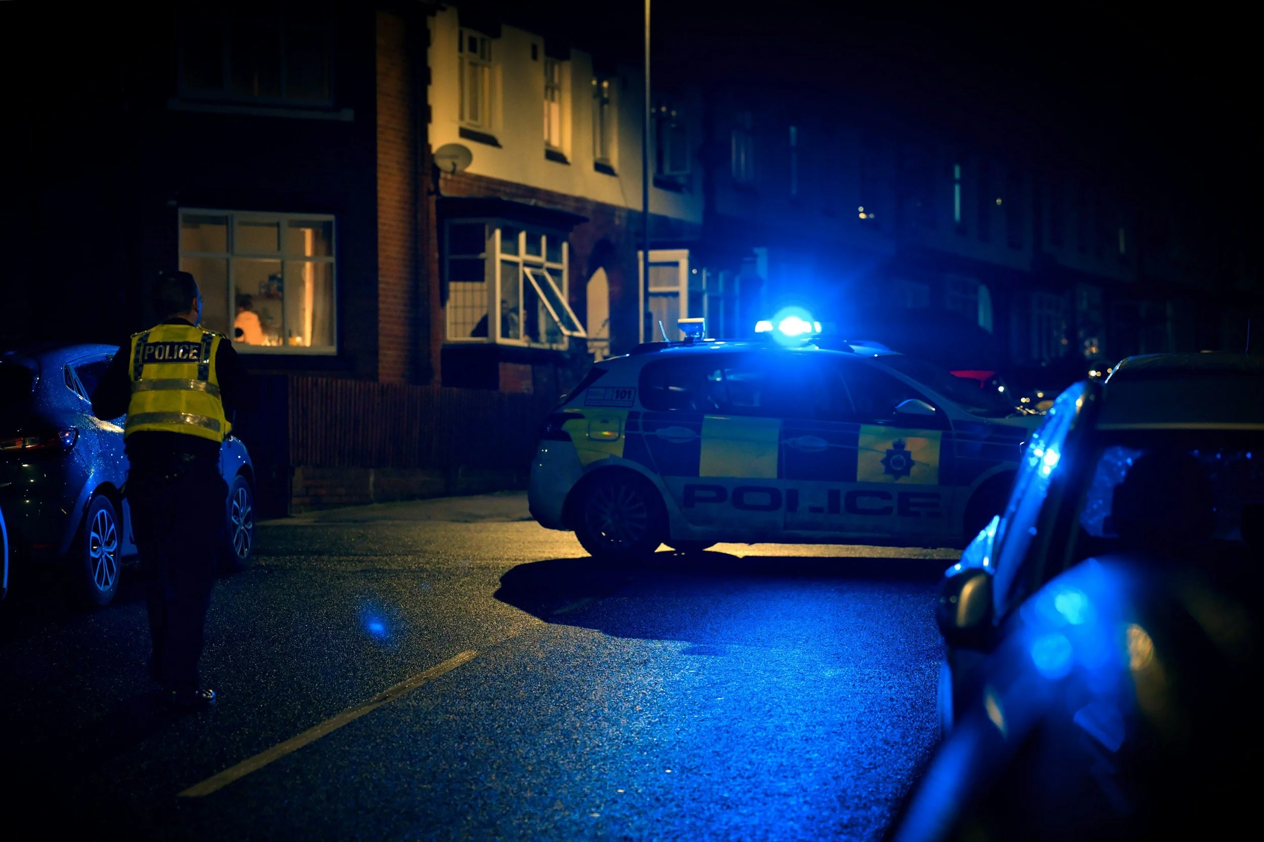 Nighttime scene of police officers and police cars with flashing lights parked on a residential street, with an officer standing near the vehicles and illuminated windows in the background.