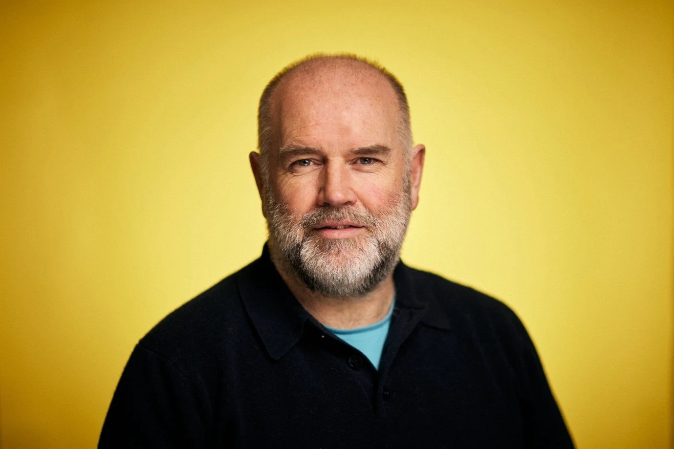 Head and shoulders portrait of a middle-aged man with a beard and bald head, wearing a black shirt against a yellow background.
