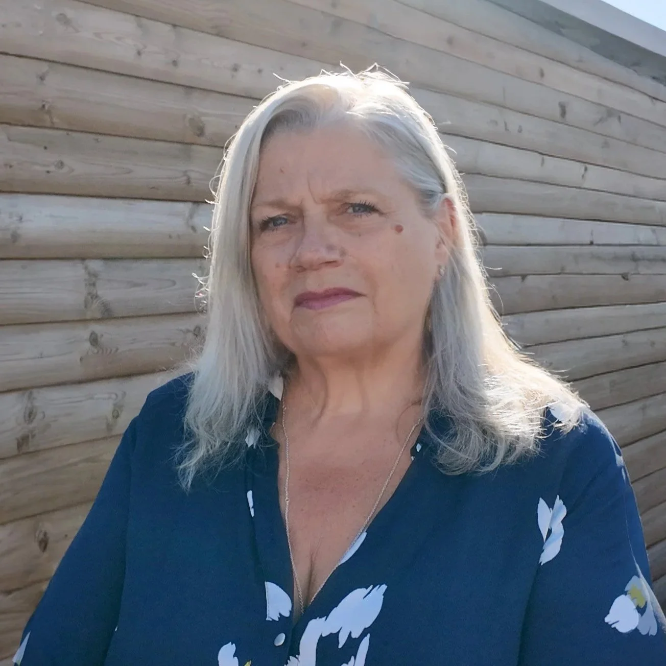 A woman with long gray hair and a serious expression, standing outdoors in front of a wooden wall, wearing a dark blue top with a floral pattern.