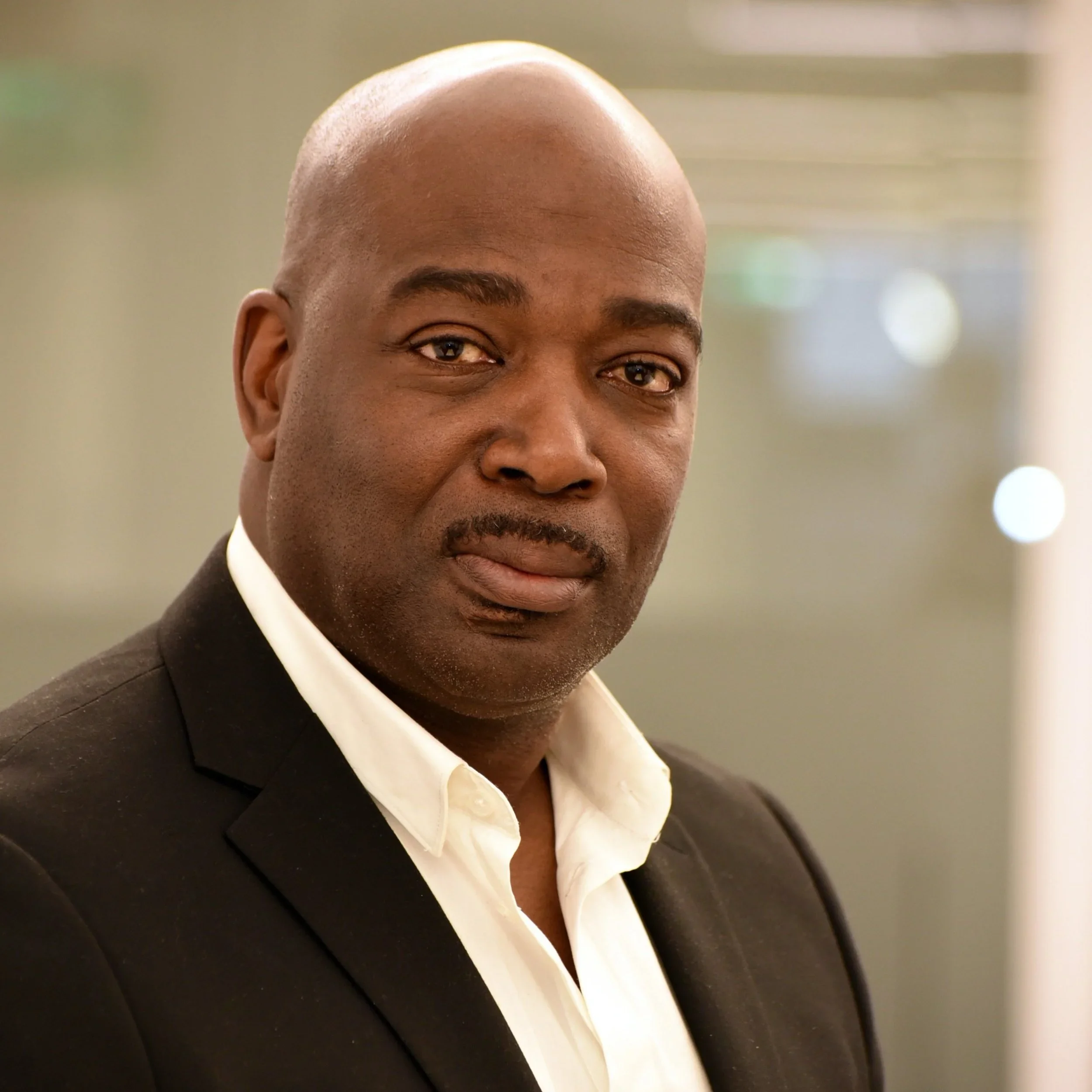 A middle-aged Black man in a black suit jacket and white shirt, looking into the camera with a neutral expression, indoor setting.