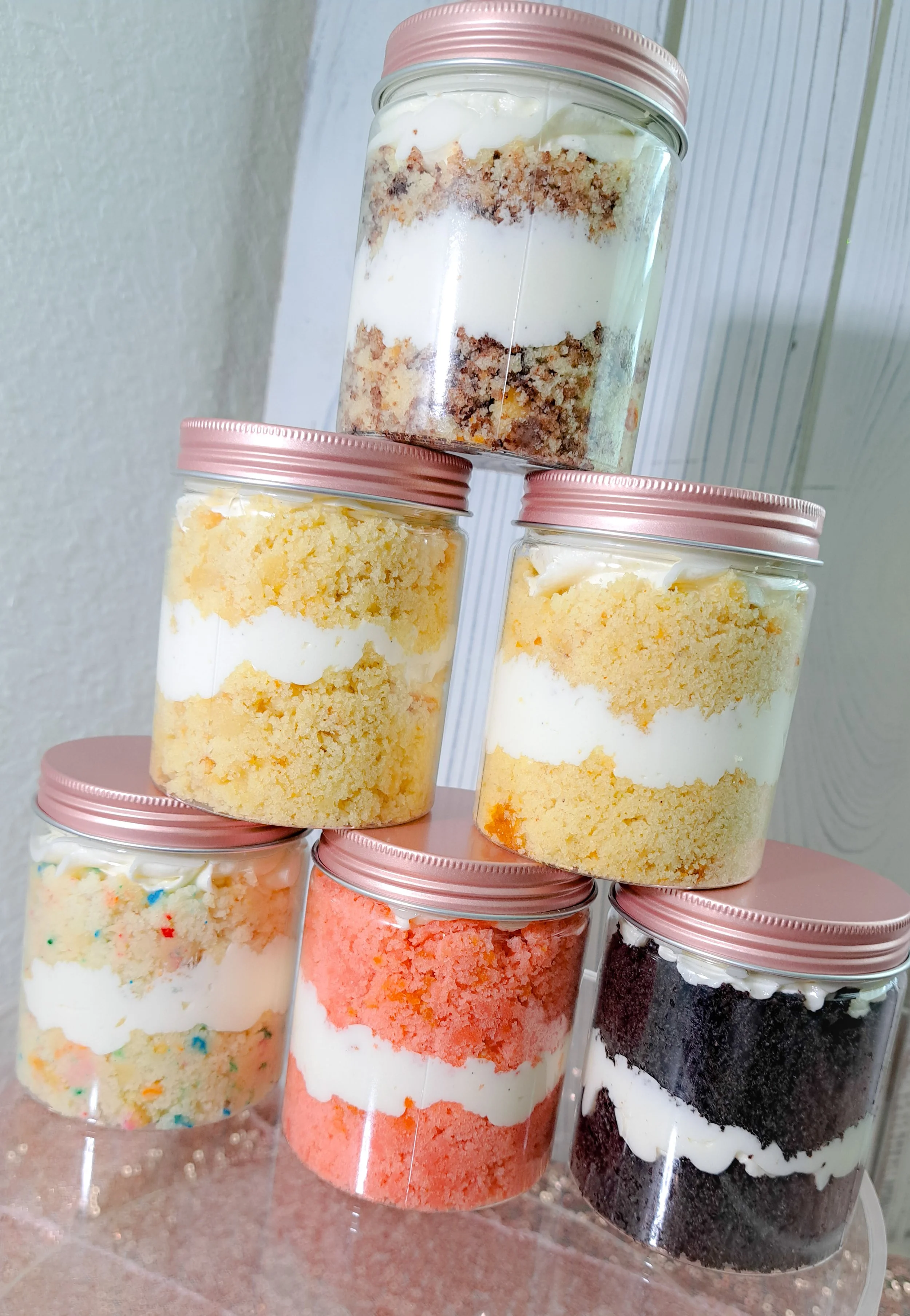 Six jars of layered cake with different colors and flavors, topped with pink lids, stacked in a pyramid on a pink surface