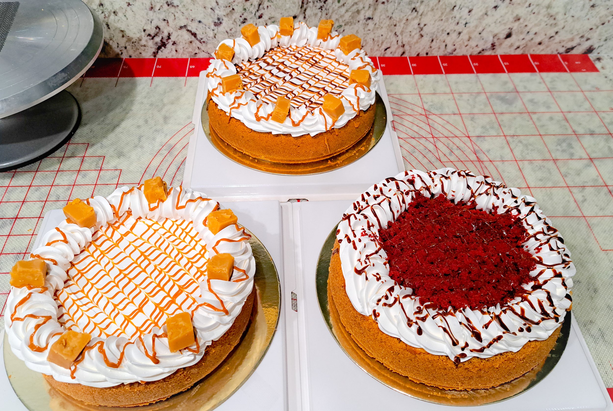 Three decorated cakes with whipped cream, caramel, and chocolate drizzle on top, placed on gold and white bases on a red and white checkered table.