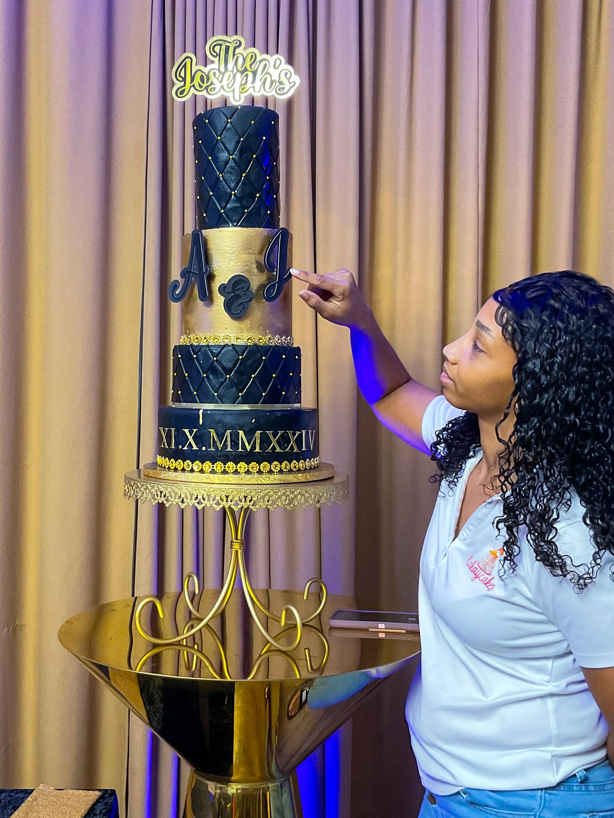 A woman with curly black hair dressed in a white shirt is pointing at a multi-tiered cake with gold and black decorations, placed on a golden stand against a beige curtain backdrop.