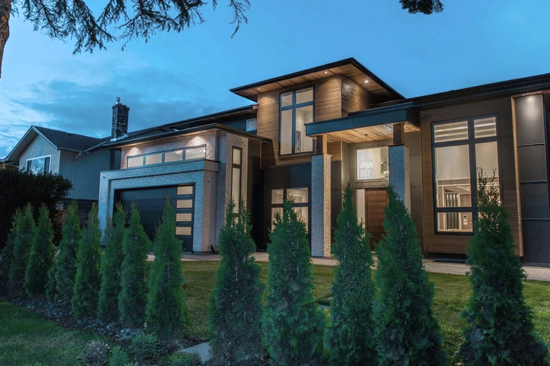 Modern house with large windows, wooden and stone exterior, front yard with shrubs, and a blue evening sky.