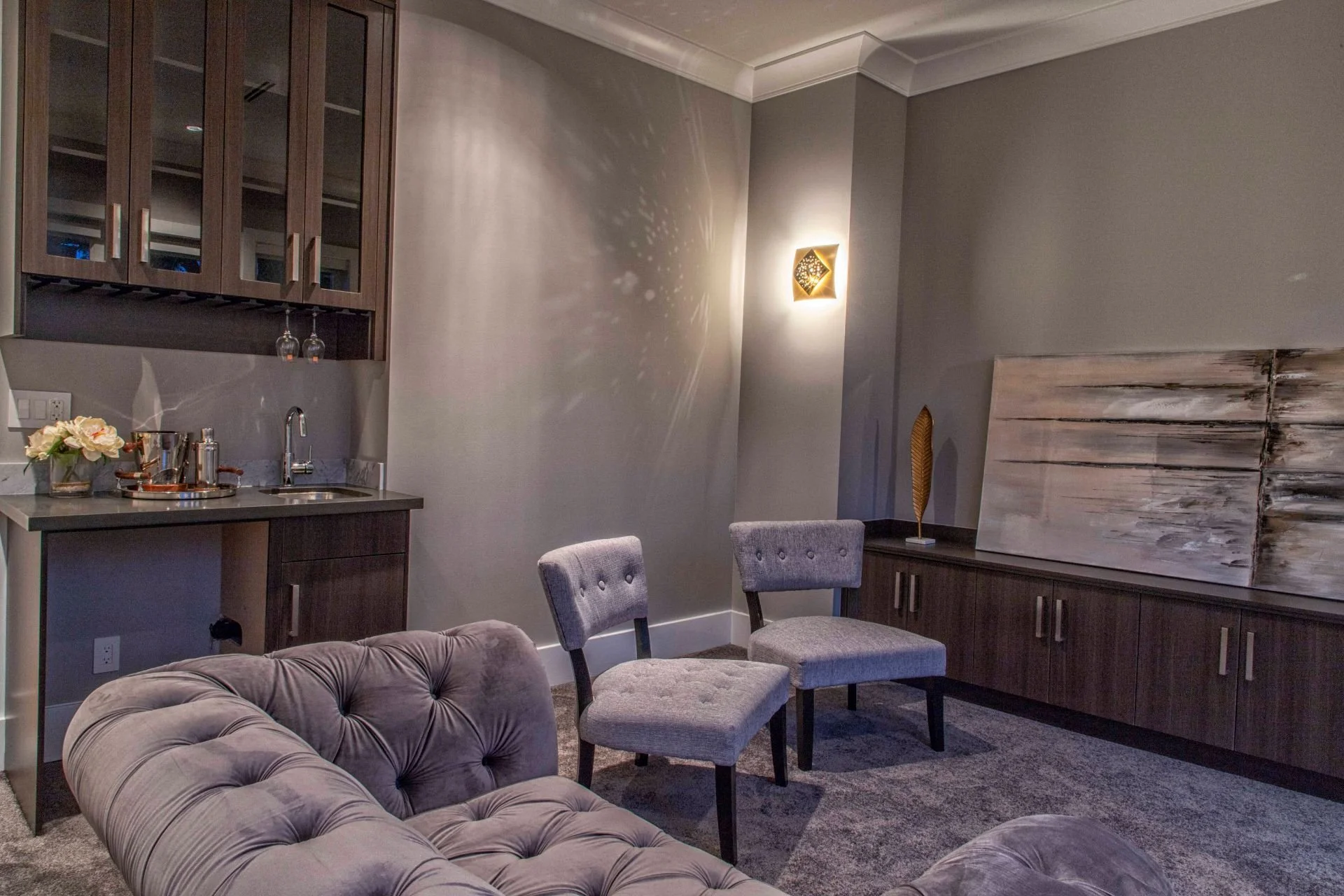A living room corner with two gray upholstered chairs, a plush gray sofa, a wooden cabinet with artwork above, and a wall-mounted light fixture casting decorative shadows.