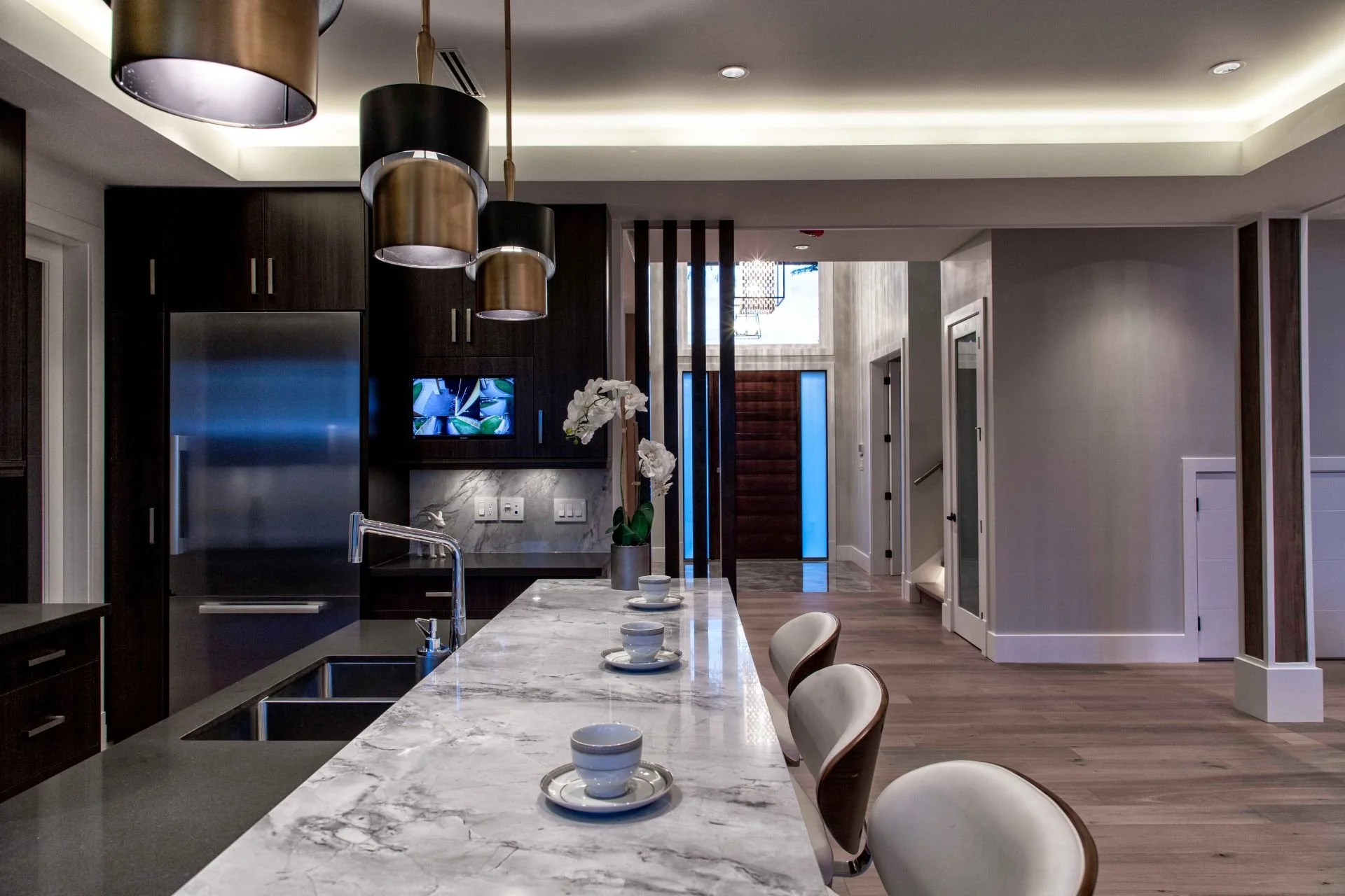 Modern kitchen with a marble island, white chairs, dark wood cabinetry, stainless steel refrigerator, centerpieces with white flowers, and pendant lights.