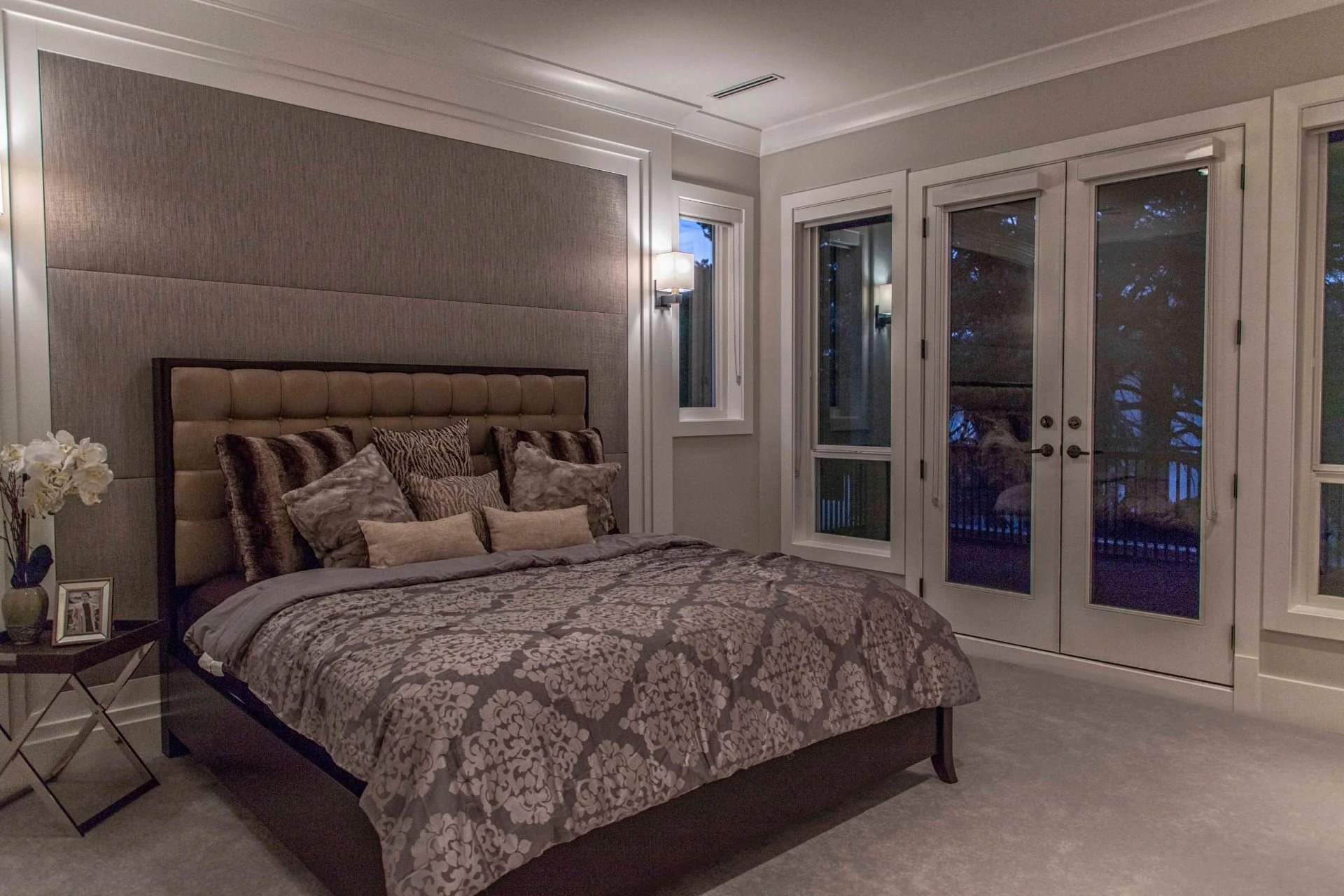 A cozy bedroom at dusk featuring a large bed with a patterned comforter and multiple pillows, a side table with a lamp, and French doors leading to a balcony, with trees visible outside.