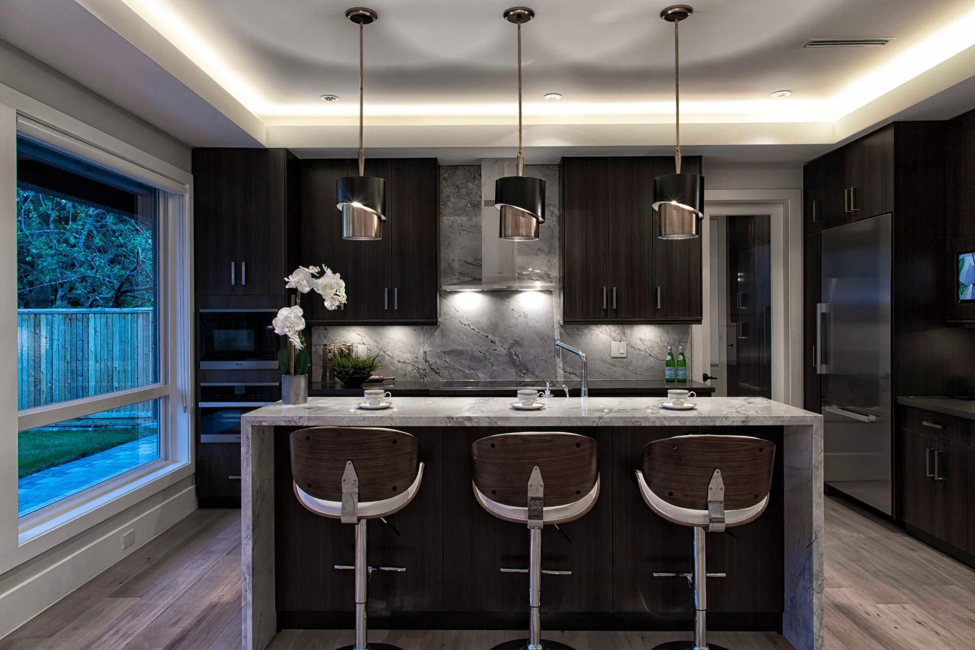 Modern kitchen with dark wood cabinets, a large marble island with three barstools, pendant lighting, and a window overlooking a backyard.