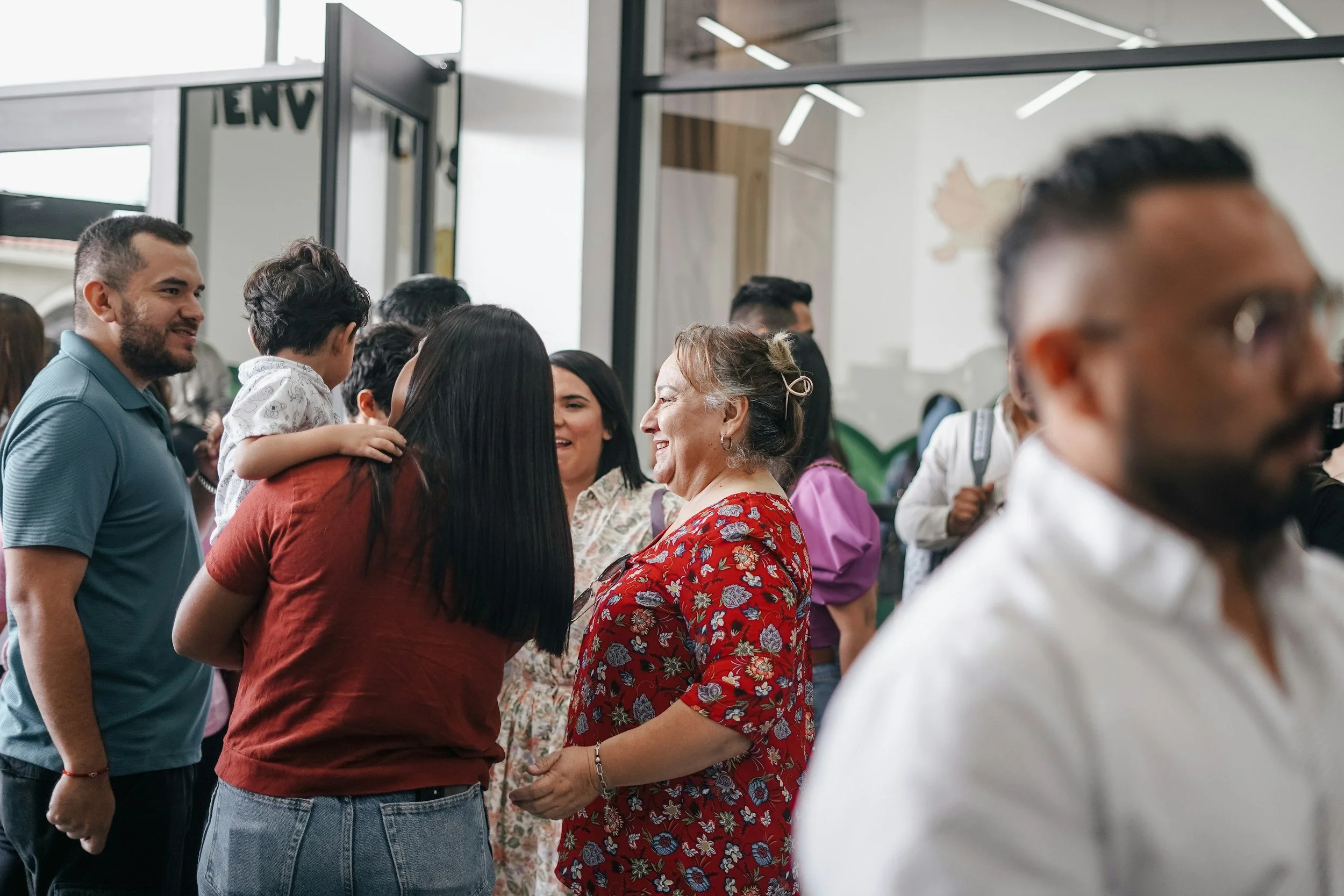 People socializing indoors, including women and a man, with a focus on a woman in a red floral top talking to a woman holding a child.