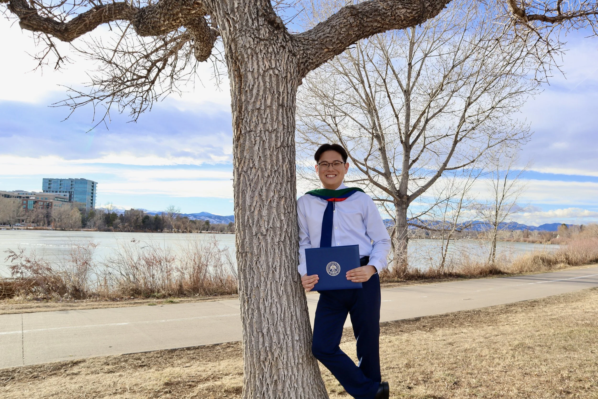 Young man in a white shirt and navy tie holding a diploma standing by a tree along a lakeside with leafless trees, mountains in the background on a partly cloudy day.