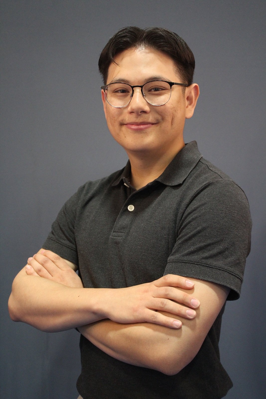 A young man wearing glasses in a dark gray polo shirt standing with arms crossed against a gray background.