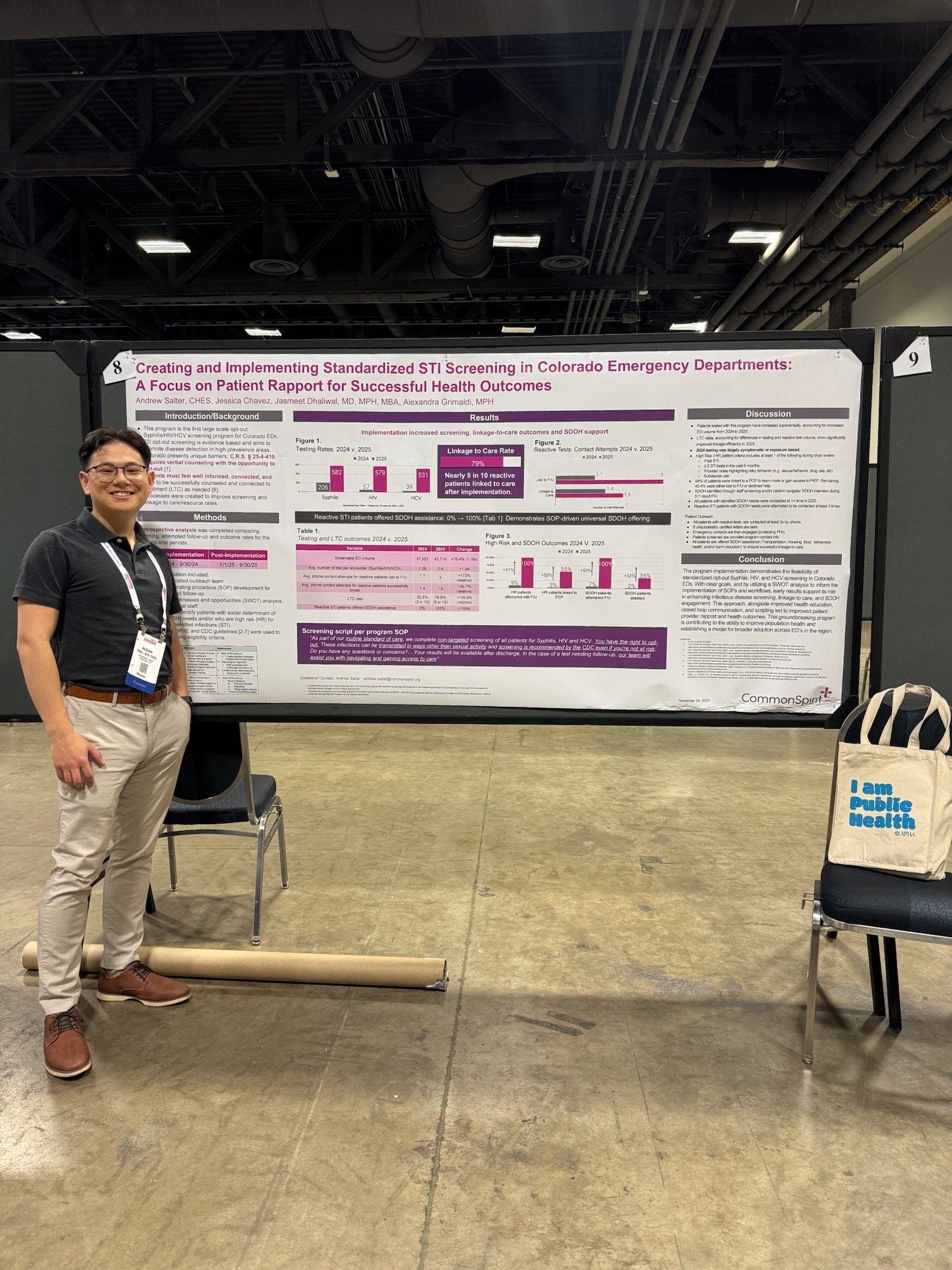 A man standing next to a scientific research poster on a display board at a conference. The poster is titled 'Creating and Implementing Standardized STI Screening in Colorado Emergency Departments: A Focus on Patient Rapport for Successful Health Outcomes.' The man is wearing glasses, a black shirt, khaki pants, and brown shoes, with a conference badge around his neck. There is a chair nearby with a tote bag that reads 'I am Public Health' and some floor covering.
