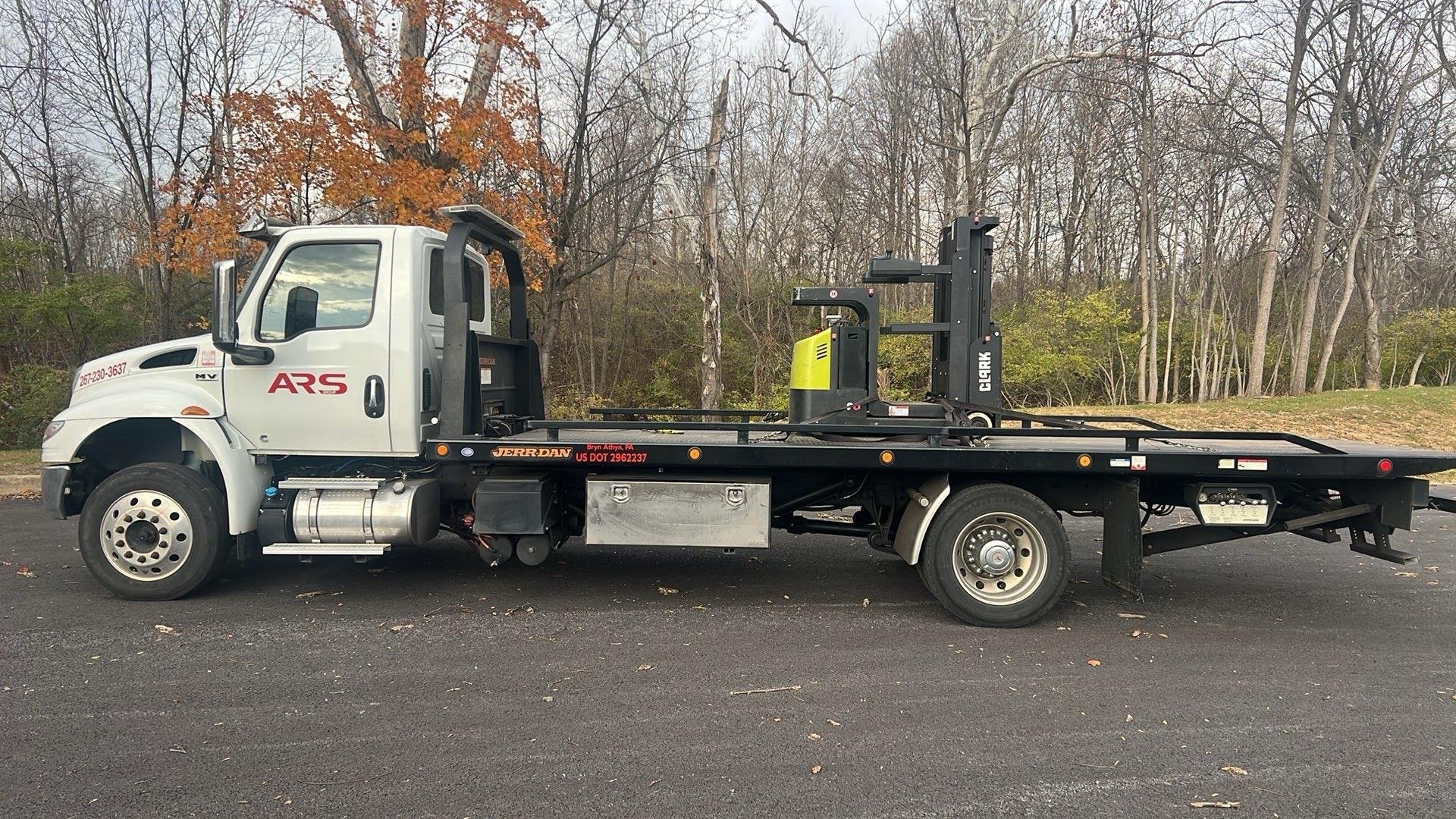 A flatbed tow truck parked on a paved surface with leafless trees and some colored foliage in the background. The truck has a white cab and a black flatbed with a yellow and black forklift attached.