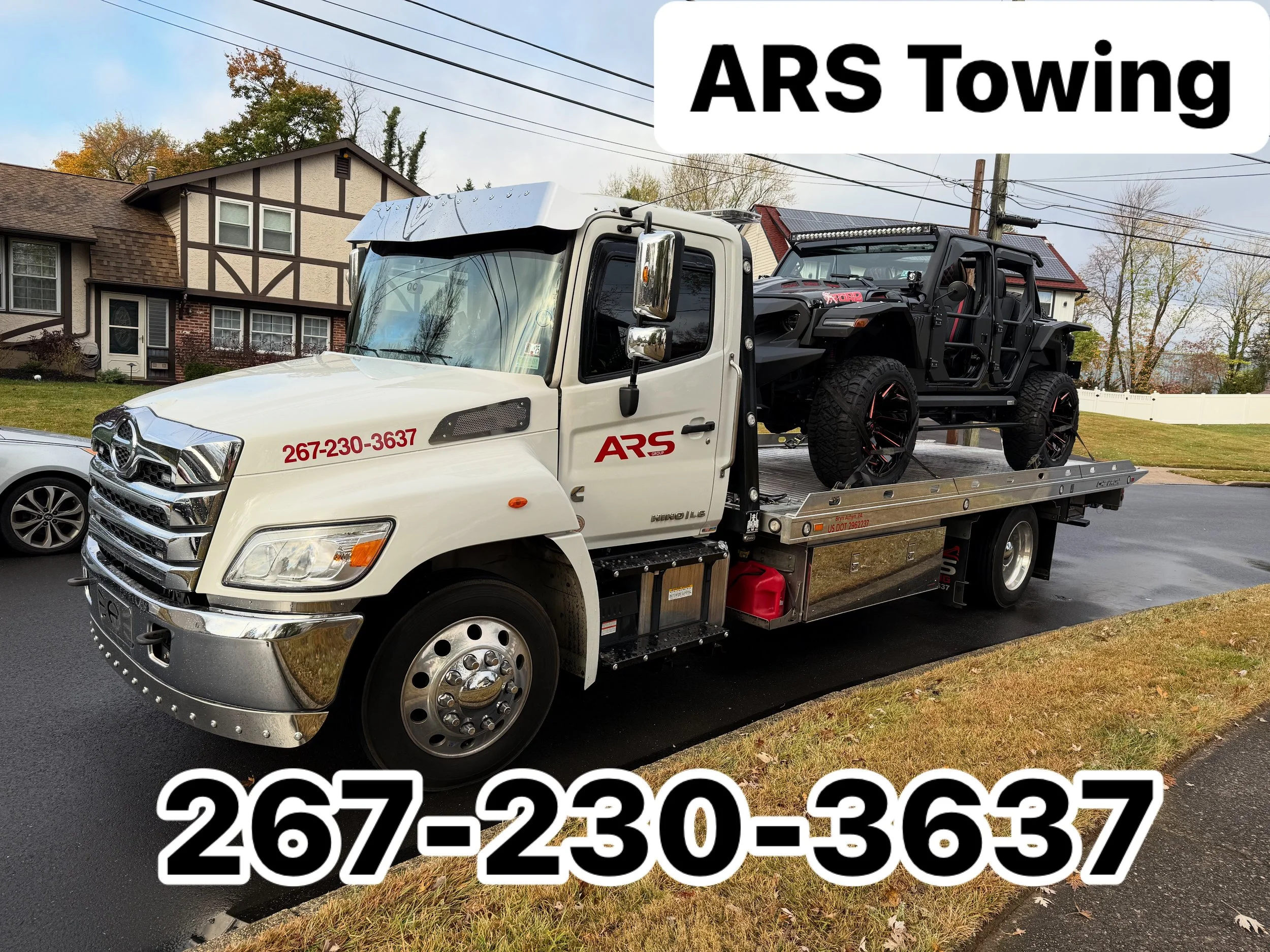 A white tow truck with ARS Towing branding and a phone number on the side, transporting a black off-road vehicle on its flatbed. The background features suburban houses, a green lawn, and overcast skies.