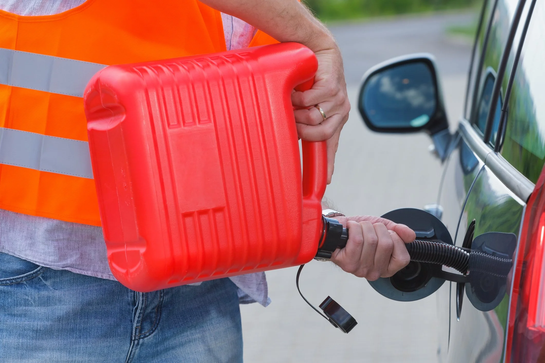Person filling a vehicle with gas from a red fuel container on the side of a road.