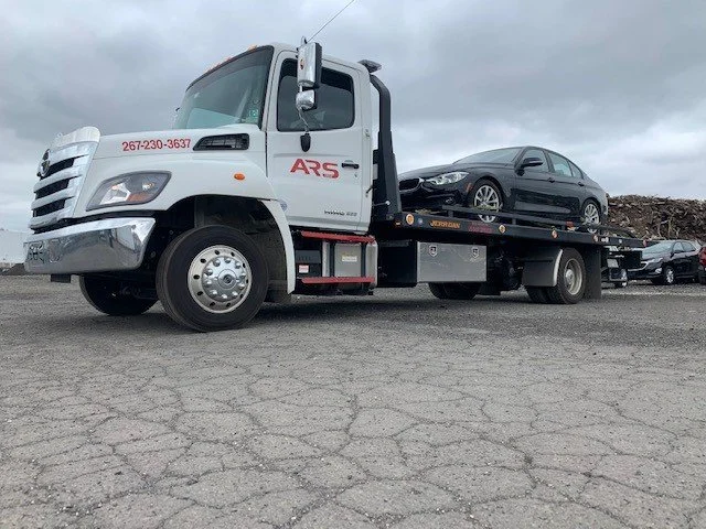 A white flatbed tow truck parked outdoors on cracked pavement, carrying a black sedan, with cloudy sky in the background.