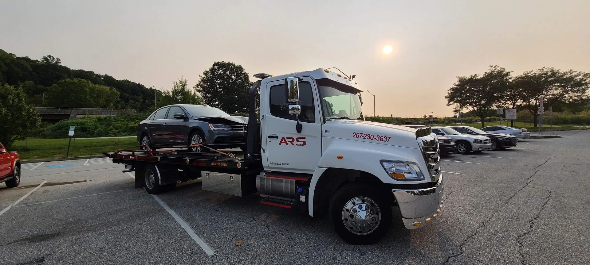 Tow truck with a black sedan on its flatbed in a parking lot during sunset.