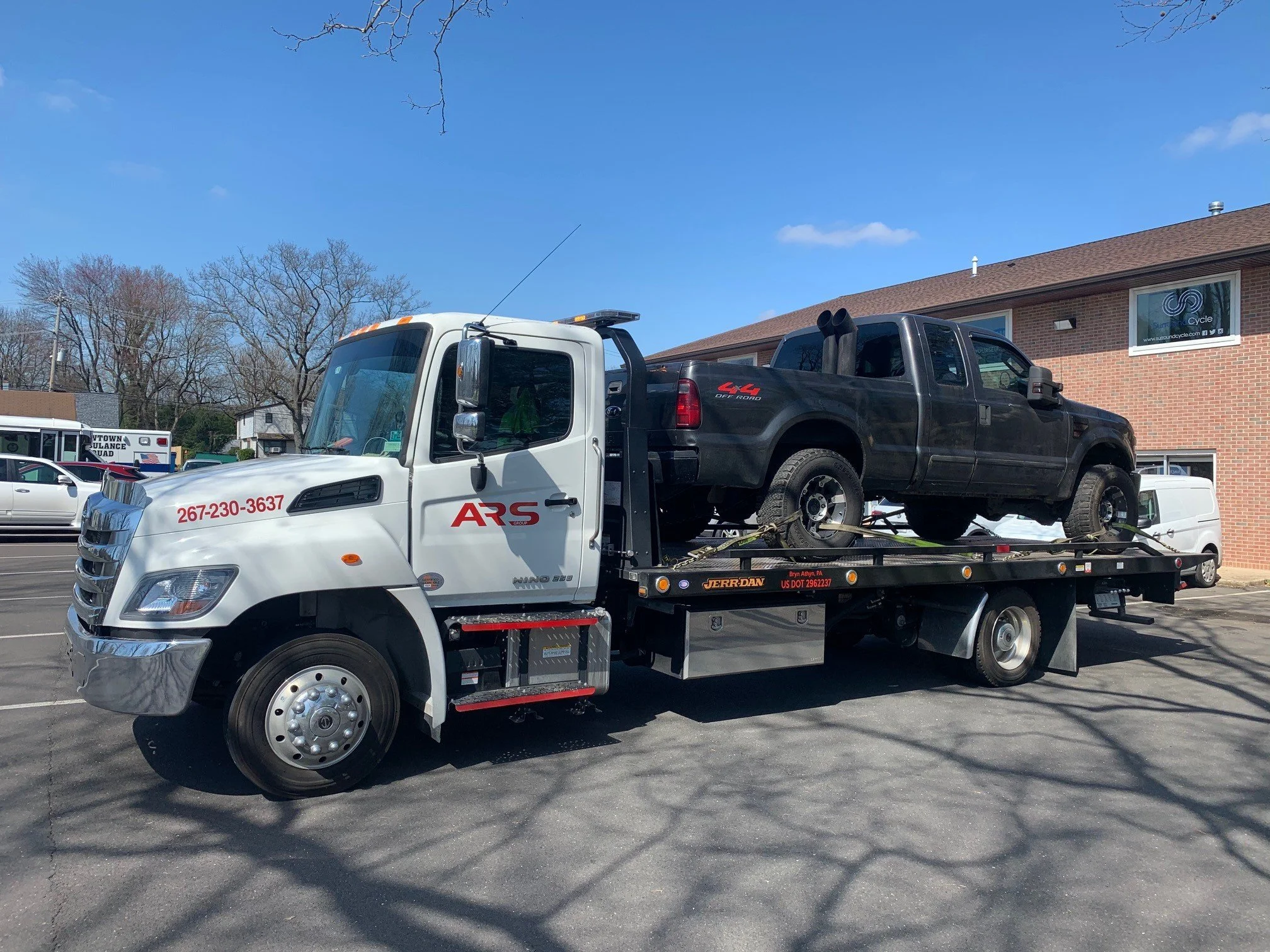 A flatbed tow truck transporting a black pickup truck on a parking lot under a blue sky with a few clouds. The tow truck has red lettering and a phone number on the side.