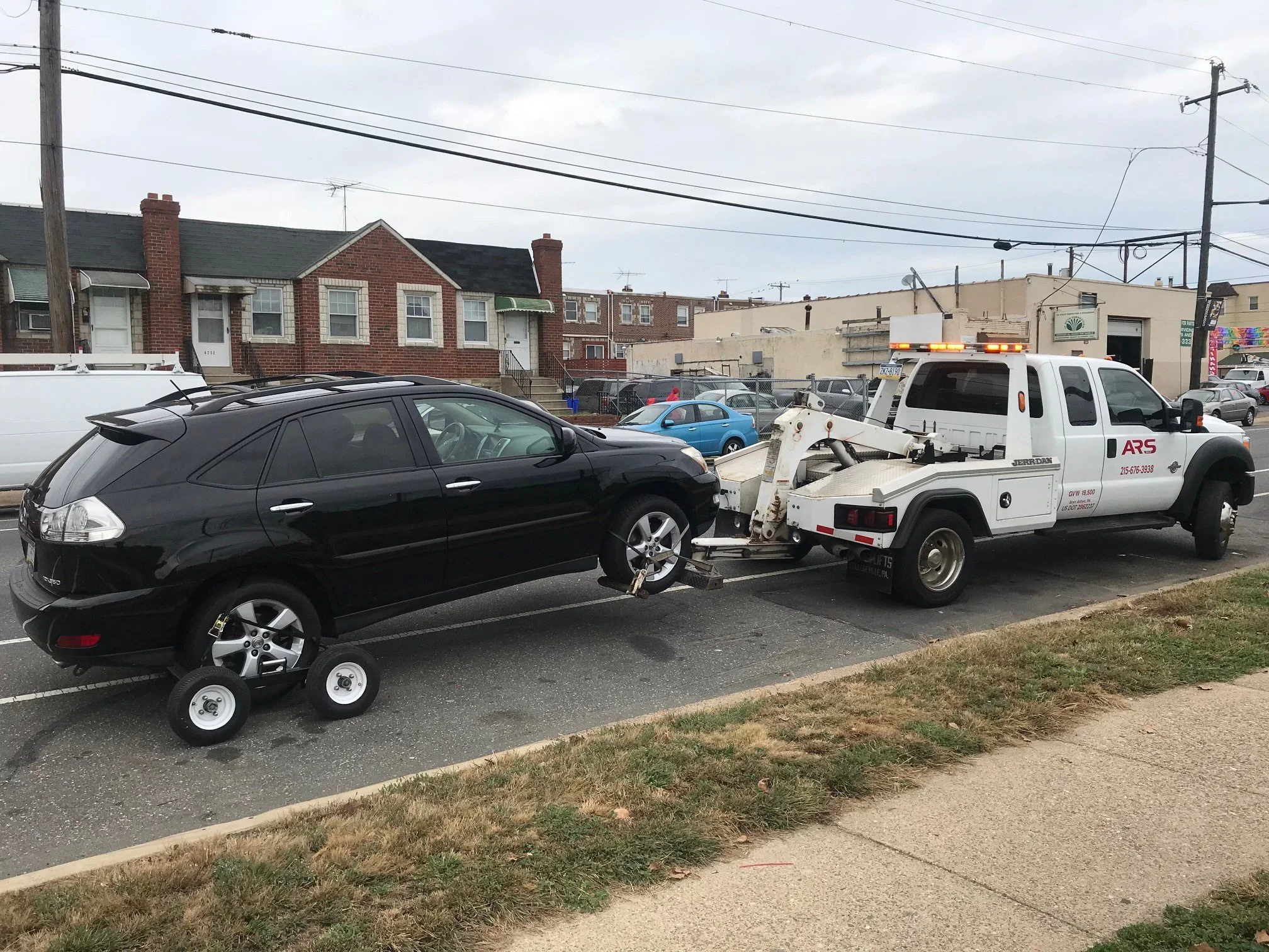A black SUV is being towed by a white tow truck in a parking lot, with a row of houses and other parked cars in the background.