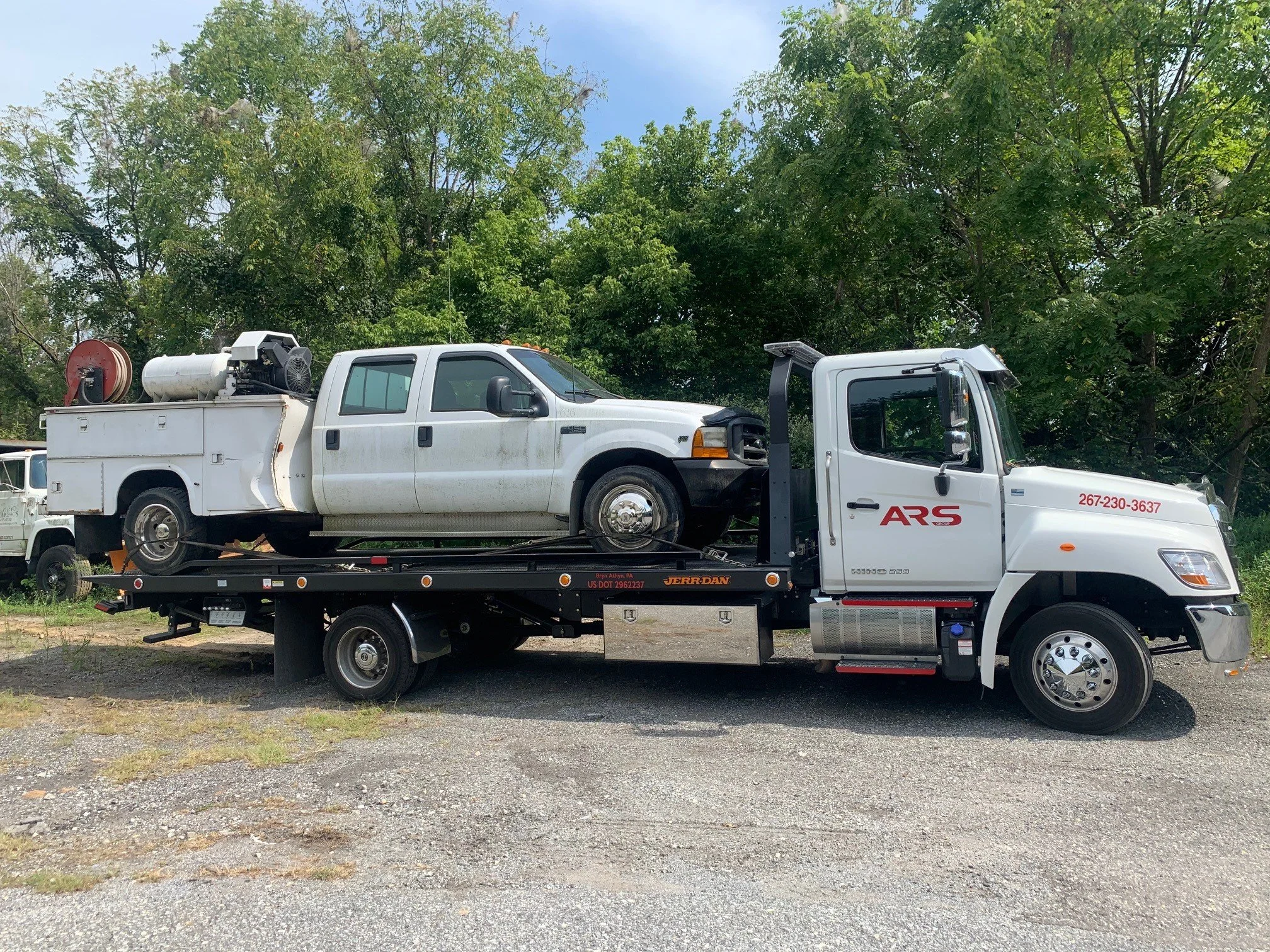 A flatbed tow truck carrying a white crew cab pickup truck with equipment on the bed, parked on a gravel surface with green trees in the background.