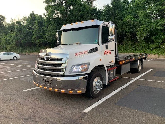 White flatbed tow truck parked in a parking lot next to trees.