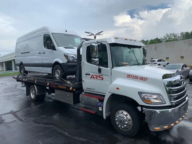 White tow truck carrying a white cargo van, parked on wet asphalt in a parking lot with other cars and a white building in the background under cloudy skies.