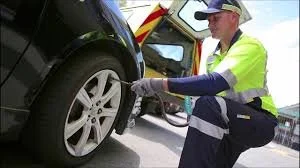 A police officer in a bright yellow vest and cap checks a black car's tire by the roadside.