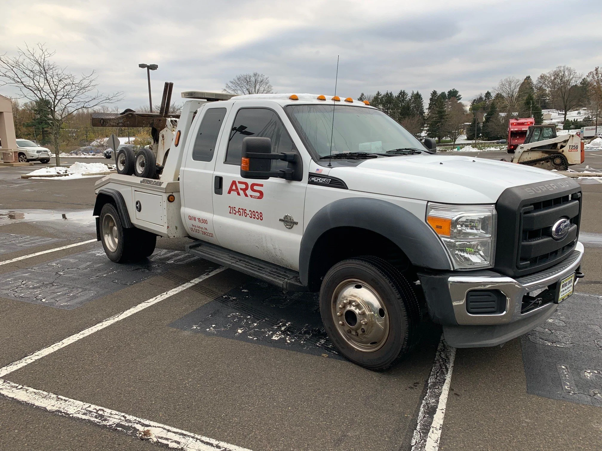 White service pickup truck with red and black ARS logo, parked in an outdoor lot with snow and trees in the background. The truck has a lift mechanism in the back.