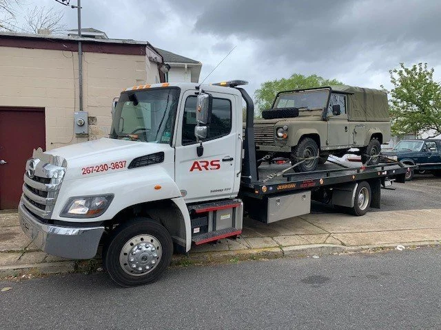 A white flatbed tow truck carrying a military-style beige vehicle with a canvas top, parked on a street in front of a building with a garage door and a cloudy sky overhead.