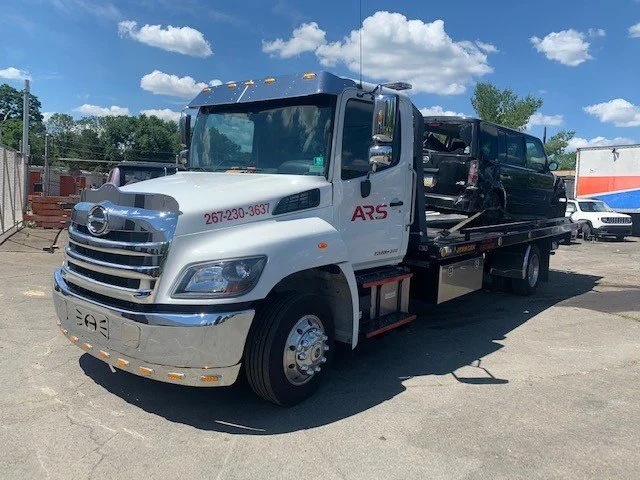 A flatbed tow truck carrying a black SUV, parked on asphalt with a blue sky and trees in the background.