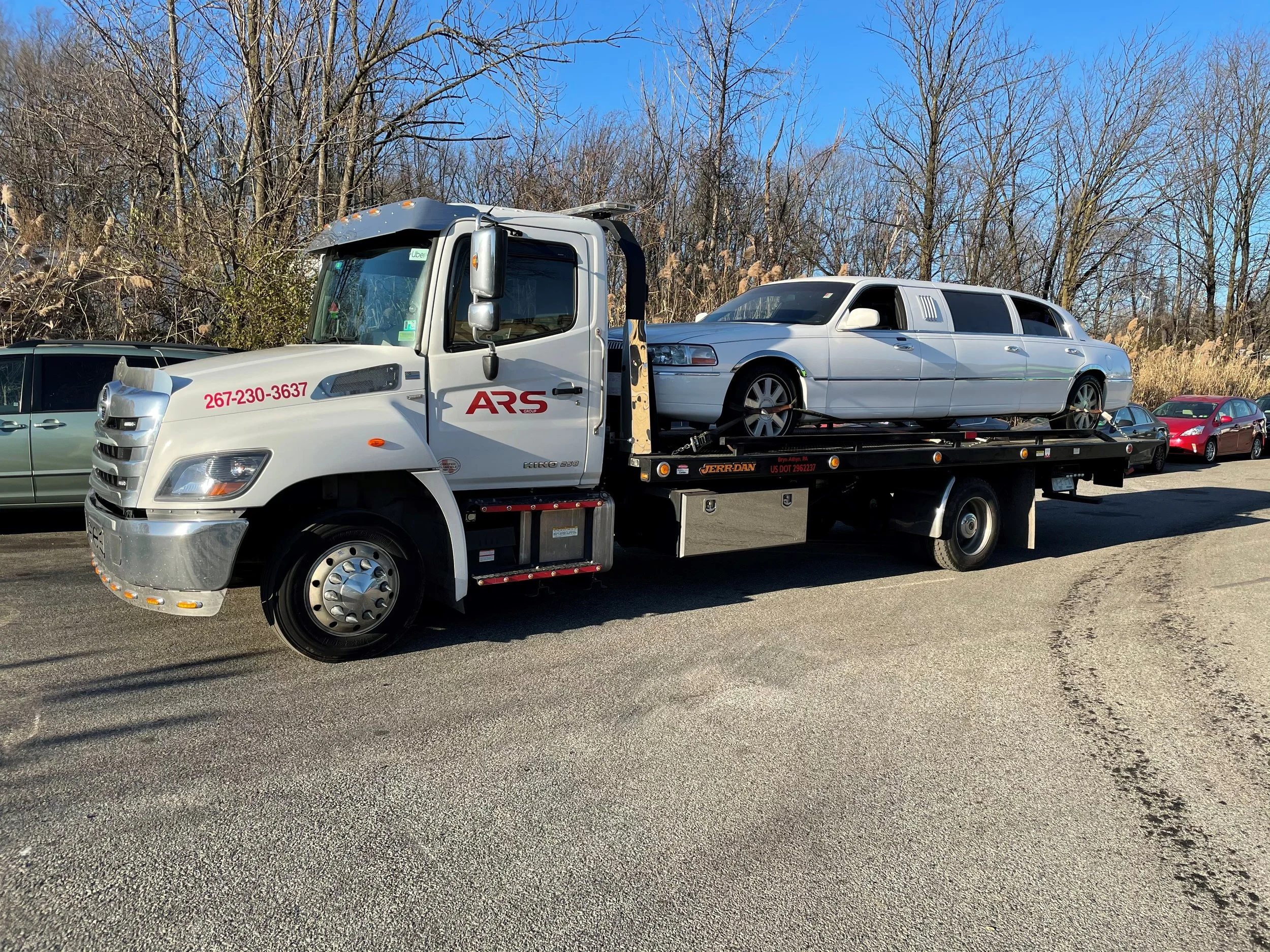 A white flatbed tow truck carrying a silver limousine on its bed, with multiple cars parked along the side of the road and leafless trees in the background.