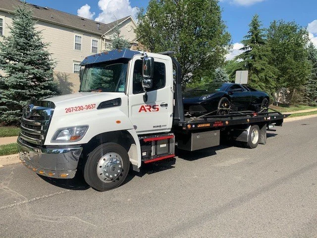 White tow truck transporting a black sports car on a flatbed in a suburban neighborhood with grass, trees, and houses.