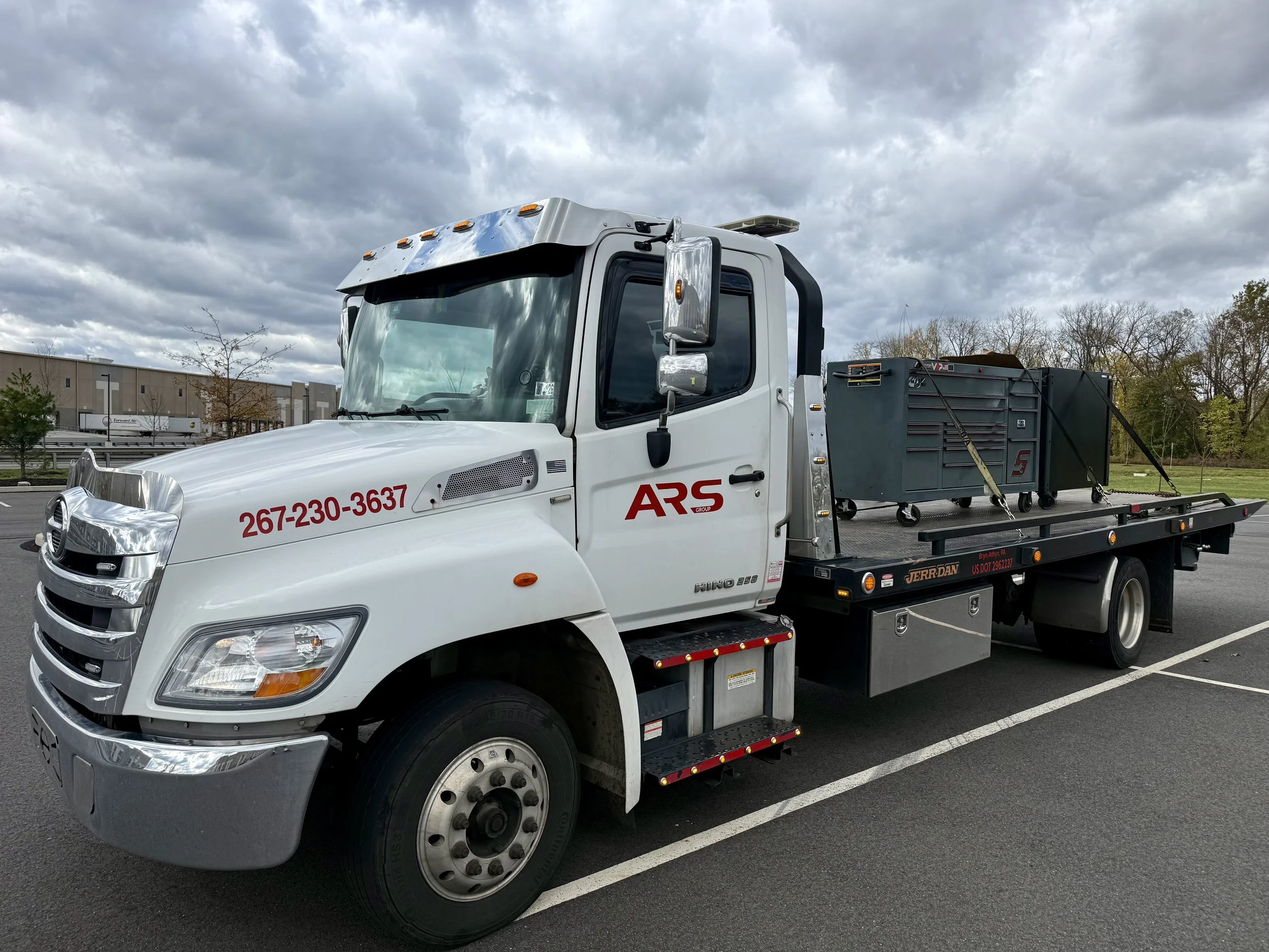 A white tow truck with ARS Group branding on the door, parked in an empty parking lot under cloudy skies. The truck is carrying a gray equipment container on its flatbed.